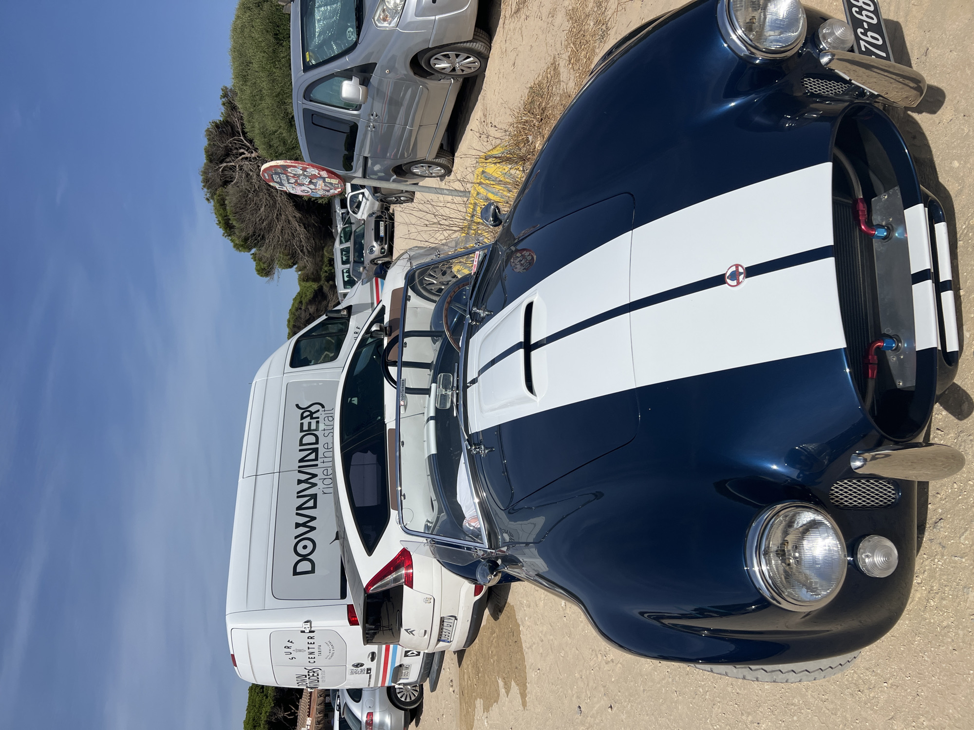 A dark blue classic convertible with white racing stripes parked on a sandy lot under a bright blue sky with scattered clouds. A white van with "DOWNWINDERS surf center" and "ride the strait" branding is parked behind the convertible. Several other vehicles, including a silver minivan and a white SUV, are visible in the background. Trees and vegetation are present on the right side of the image. The license plate of the convertible reads "76-68". The scene appears to be outdoors, possibly a parking area near a beach or recreational facility in Tarifa, Spain, given the branding on the van.