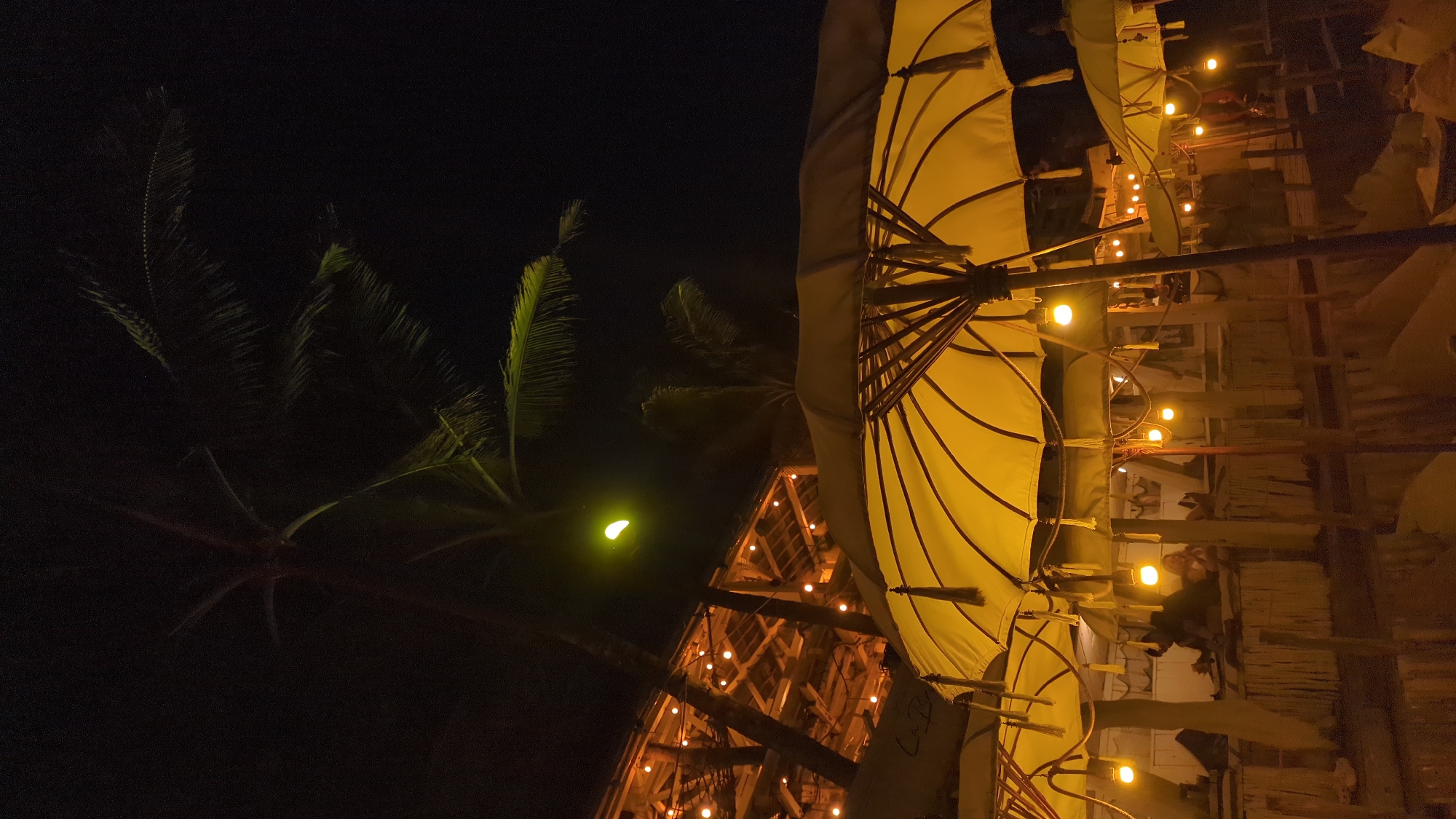 At night in North Kuta, Indonesia, a low-angle shot captures a scene illuminated by yellow light. In the foreground, a row of light-colored umbrellas are opened, each illuminated by string lights. Behind the umbrellas, a wooden structure is visible, also lit with string lights. In the background, the silhouette of a palm tree is against the dark sky. The moon is a small, crescent shape in the sky.