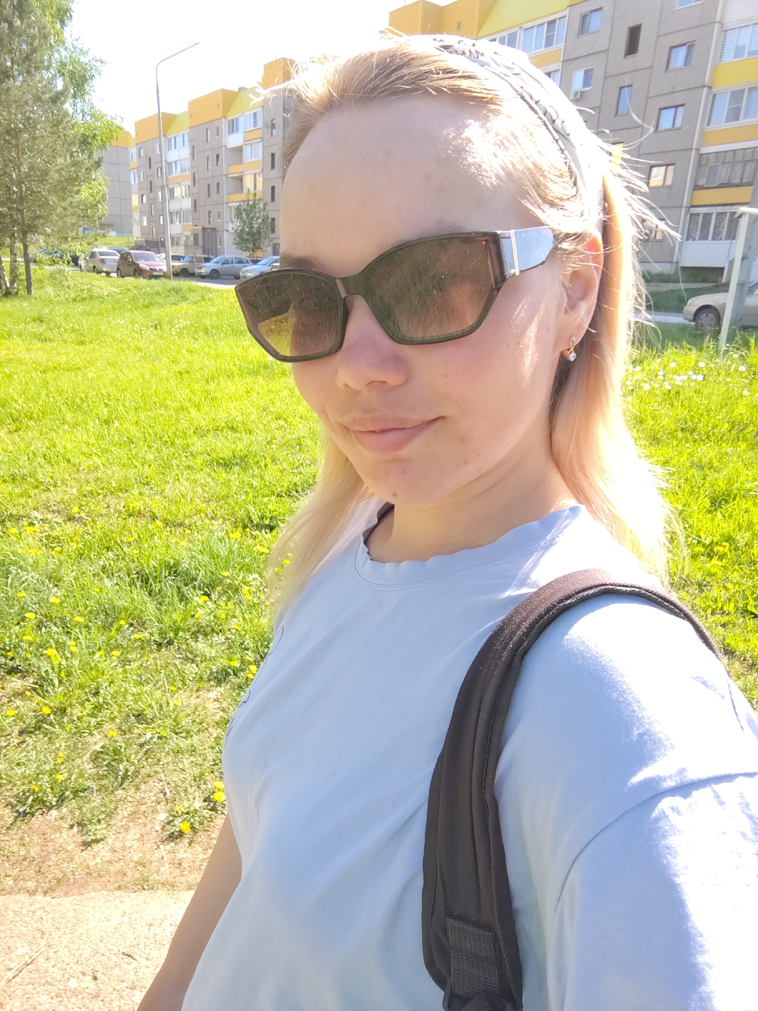 A woman wearing sunglasses and a backpack stands on a grassy lawn next to a sidewalk in the urban area of Марковский, Russia. The woman has blonde hair with a white bandana. In the background, there is a light post and a gray and yellow apartment building, along with parked cars and trees.