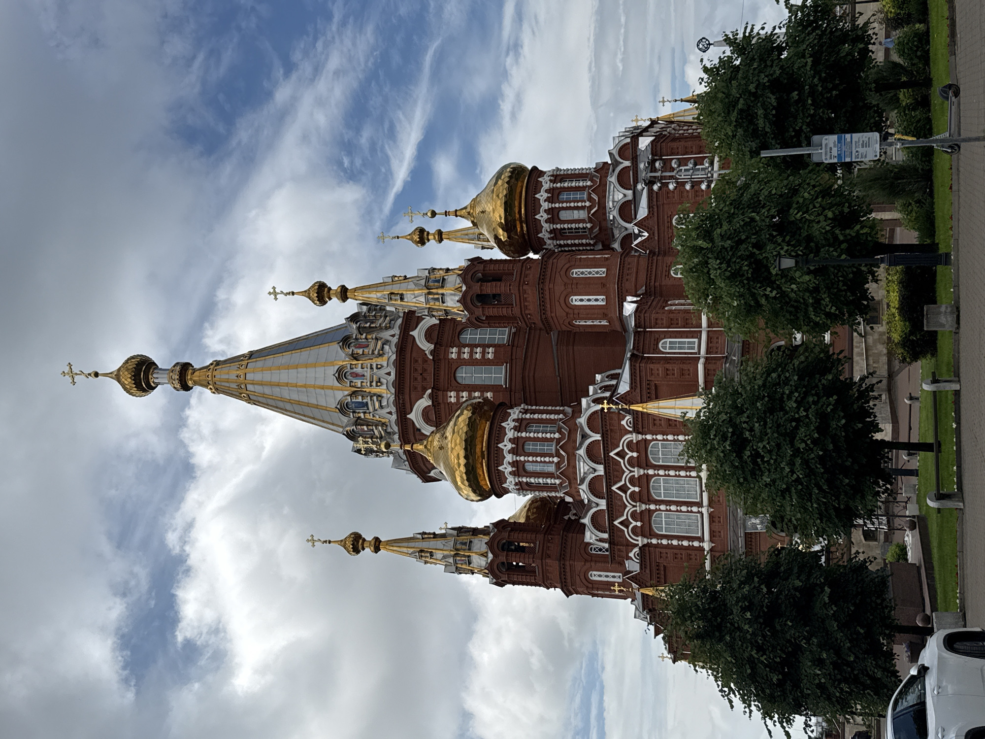 The image shows the Holy Trinity Cathedral in Izhevsk, Russia, a large, ornate red brick church with multiple golden domes topped with crosses. The cathedral is set against a cloudy sky with patches of blue. In the foreground, there are several green trees, a white car, a street light, a bench, and a scooter parked near a signpost. The cathedral's architecture features white decorative elements and numerous windows. The overall scene depicts a religious landmark in an urban setting.