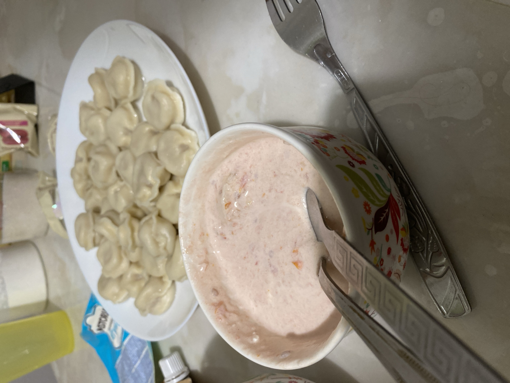 This indoor photograph, taken in Chaikovsky, Russia, showcases a meal consisting of dumplings and a creamy sauce. The main subjects are a white oval plate heaped with numerous light-colored, cooked dumplings (resembling pelmeni or vareniki) and a ceramic bowl containing a pale pink, creamy sauce with visible reddish specks, possibly a flavored sour cream. A pair of silver tongs, adorned with a Greek key pattern on its handles, is placed within the sauce, while a silver fork with an ornate handle lies on the countertop next to the bowl.
The scene appears to be set on a light-colored, possibly off-white, countertop or table surface, which shows some minor smudges or stains. In the background, various household items are partially visible and out of focus, including a white cylindrical roll (likely paper towels), a bright yellow container, a small white bottle with a ribbed cap, and a blue package on which the partial word "VICTORY" can be discerned. The lighting suggests an artificial indoor environment. The image captures a casual, ready-to-eat meal. No people are visible, and no specific time of day or weather can be inferred beyond the indoor setting.