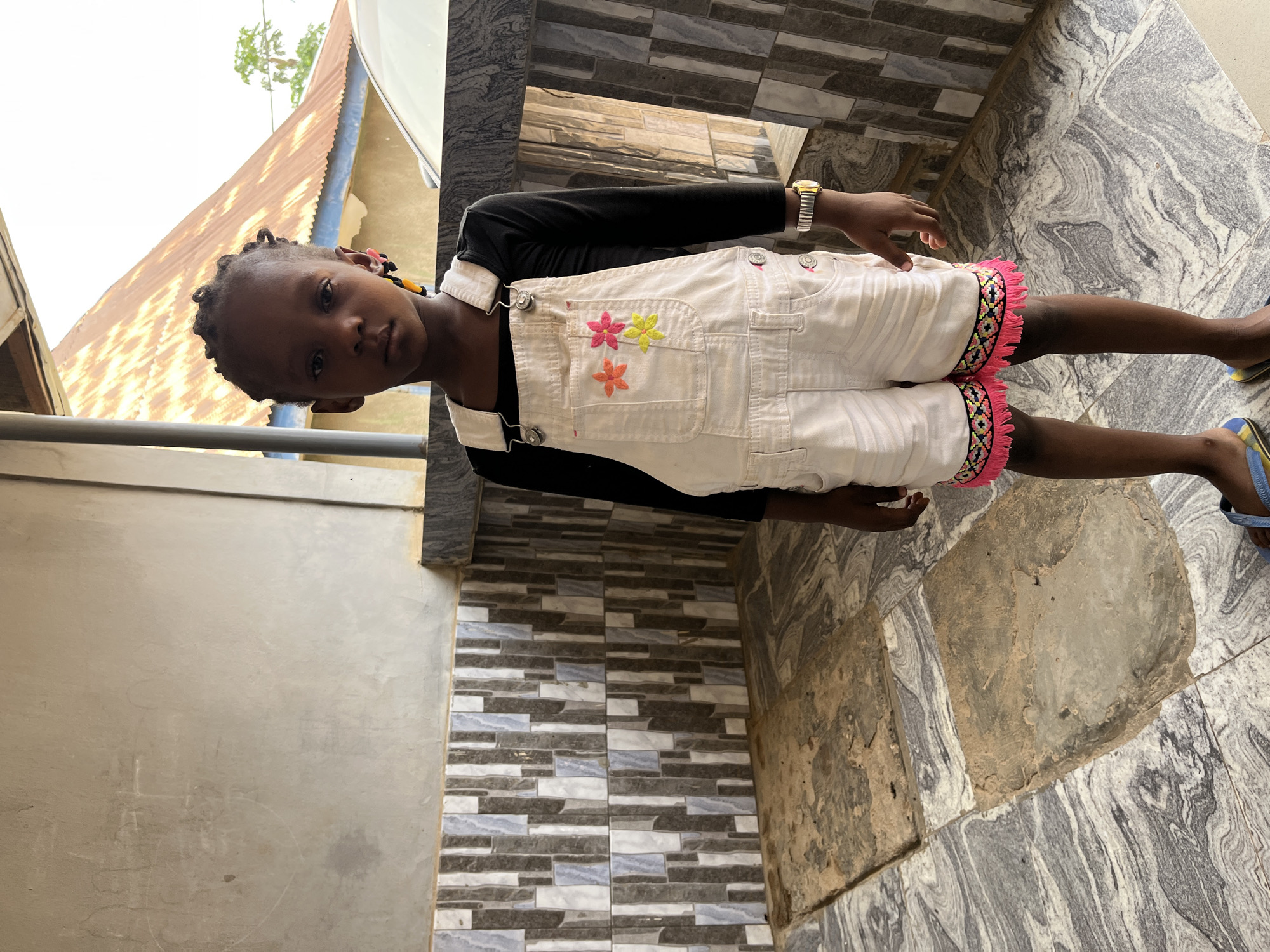 A young girl stands on a marble patterned tile floor. She wears a black long-sleeved shirt under white overalls with pink, yellow, and orange flower appliques on the bib pocket, and pink, black, and white fringe at the bottom of the shorts. She also wears a gold-colored watch on her left wrist and blue flip-flops on her feet. The girl is looking towards the camera with a serious expression. Her hair is braided with pink and yellow beads. In the background, there is a wall with multi-colored rectangular tiles, a grey wall, and a corrugated metal roof. The location is Madalla, Nigeria.