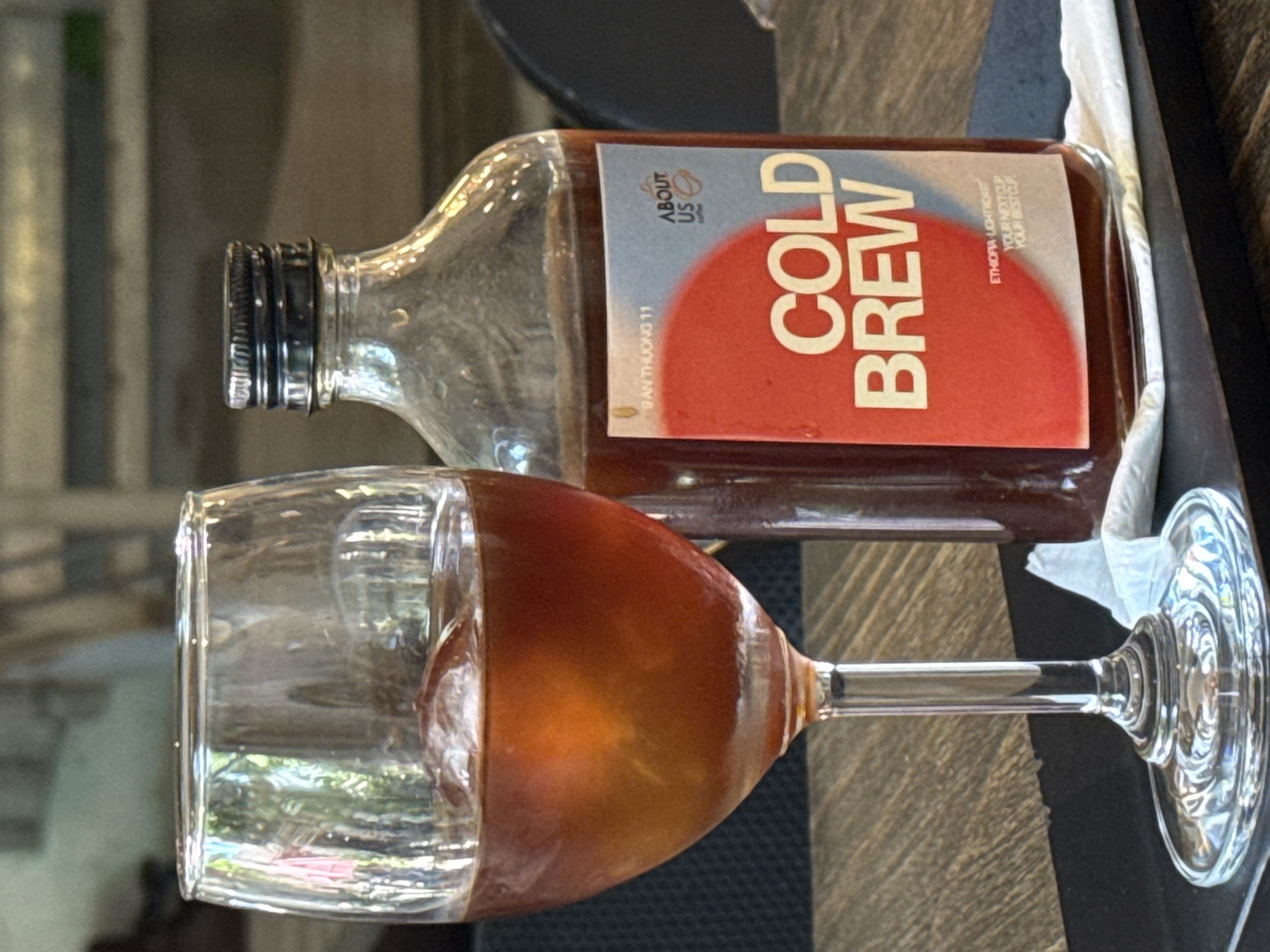 A glass of cold brew coffee with ice sits on a wooden table next to a bottled cold brew coffee. The bottle has a red and blue label that reads "COLD BREW" and "ABOUT US COFFEE". The background is blurred and appears to be an indoor setting. The overall scene suggests a cafe or coffee shop.