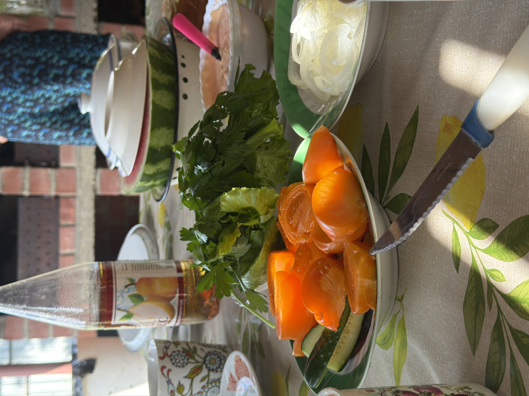 The image depicts a picnic table laden with food and drinks, set outdoors, possibly in the city of Kurgan, Russia, given the context. In the foreground, a plate of sliced orange tomatoes and cucumber is visible, alongside a bowl of sliced onions. A knife with a serrated blade and a blue and white handle rests on the patterned tablecloth. Further back, a bottle of amber-colored liquid, likely a beverage, stands near a cup with a floral design. A bunch of fresh green lettuce or herbs is also present. In the background, a watermelon is partially visible, topped with white bowls. The overall setting suggests a casual outdoor meal or gathering. A person, dressed in a blue and black patterned garment, is partially visible in the upper right portion of the frame. The environment appears to be a backyard or garden area, with a brick structure in the background. The sunlight casts shadows across the scene, indicating it is daytime.