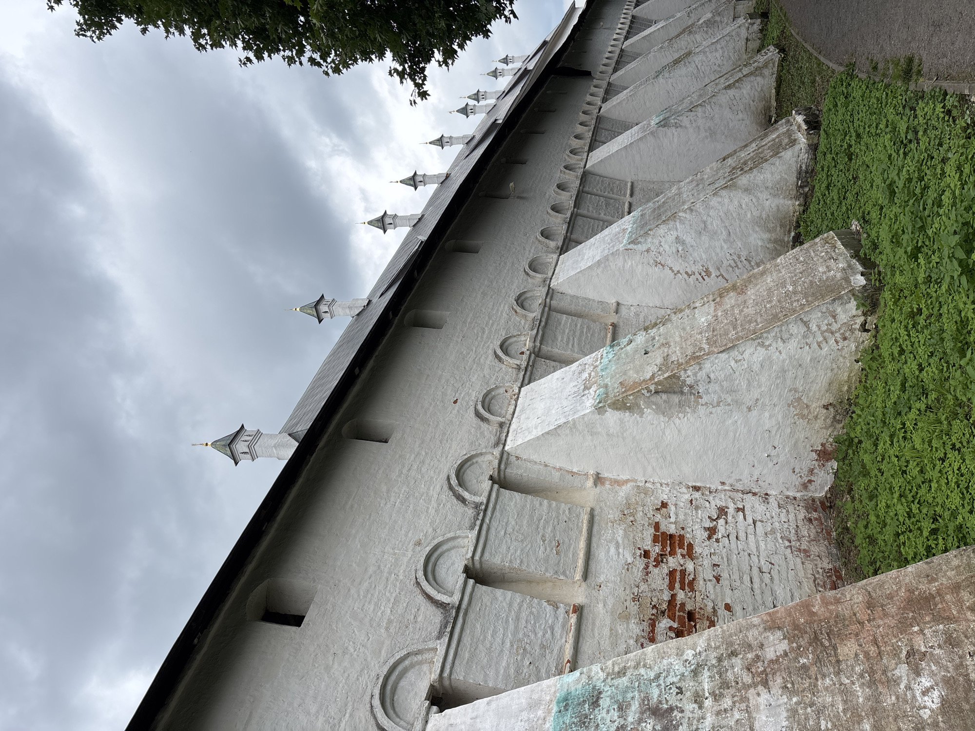 The image shows a long, white, brick wall of a religious complex, likely the Savvino-Storozhevsky Monastery in Zvenigorod, Russia. The wall is reinforced with numerous triangular buttresses, some of which show signs of wear and brickwork. Above the wall, a row of small, decorative towers with pointed roofs and golden finials rise against a cloudy, gray sky. Green foliage grows along the base of the wall and a paved path runs alongside it. A dark green tree branches into the upper right corner of the frame. There are no people or animals visible in the scene.