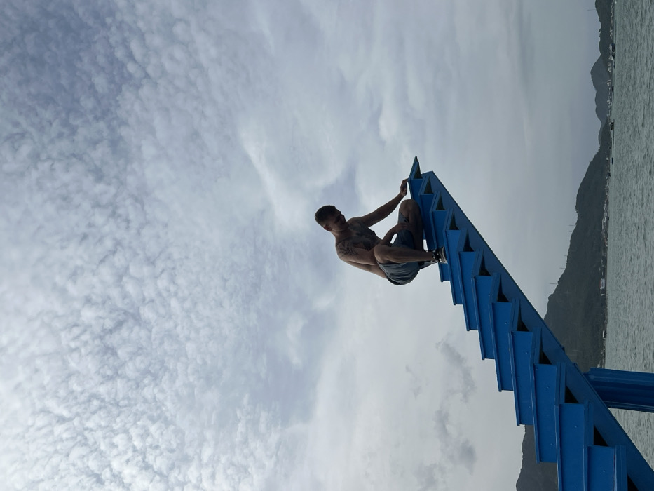 A single adult male, shirtless and visibly tattooed on his arms and chest, is depicted squatting on the top step of a prominent, bright blue, cantilevered staircase. He is wearing dark shorts and athletic shoes, and his gaze is directed downwards and slightly to his left. His right hand rests on the top step, and his left hand is on his thigh. The blue staircase, featuring approximately eight visible steps, ascends from the lower-left portion of the frame and terminates abruptly mid-air over a body of water. This scene is located in Ninh Ích, Vietnam. In the background, across the water, a distant mountainous coastline is visible, dotted with scattered low-rise structures and several floating platforms or small boats on the water near the shore. The upper portion of the image is dominated by a sky filled with a dense, textured layer of altocumulus clouds.