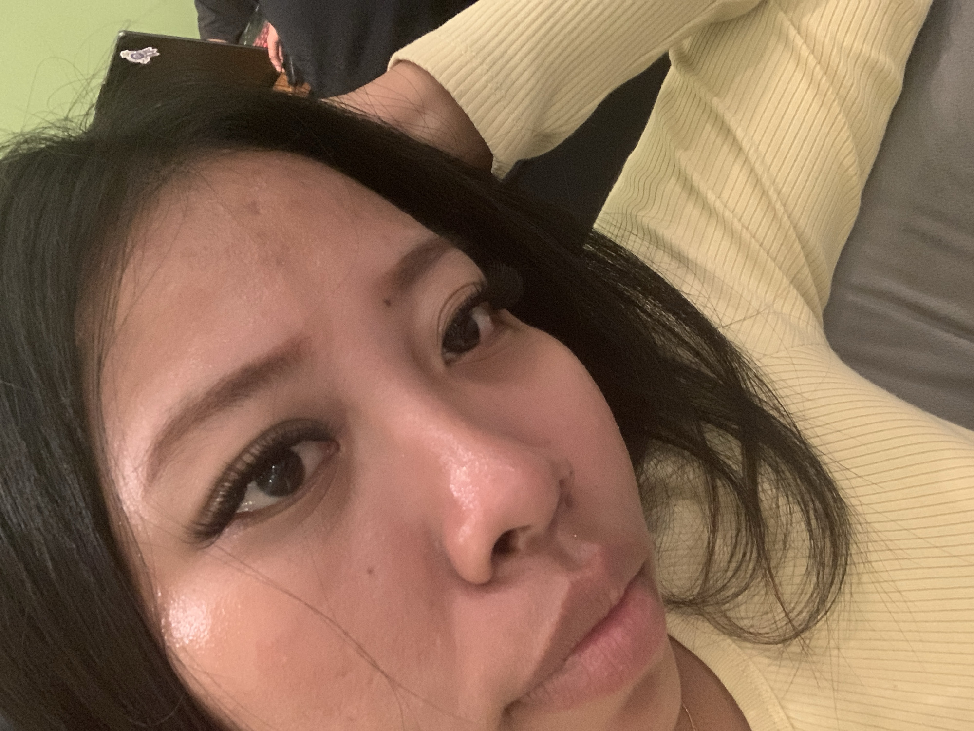 A close-up, low-angle, selfie-style photograph shows a young East Asian woman in the foreground. She has long, dark hair, and is wearing a light yellow, ribbed sweater. Her face is tilted, and her left hand is resting on her head. The background is out of focus and appears to be an indoor setting with a green wall and a laptop with a sticker on it. The lighting is soft and highlights the sheen on her skin. The image does not contain enough information to determine its location in Denpasar, Indonesia.
