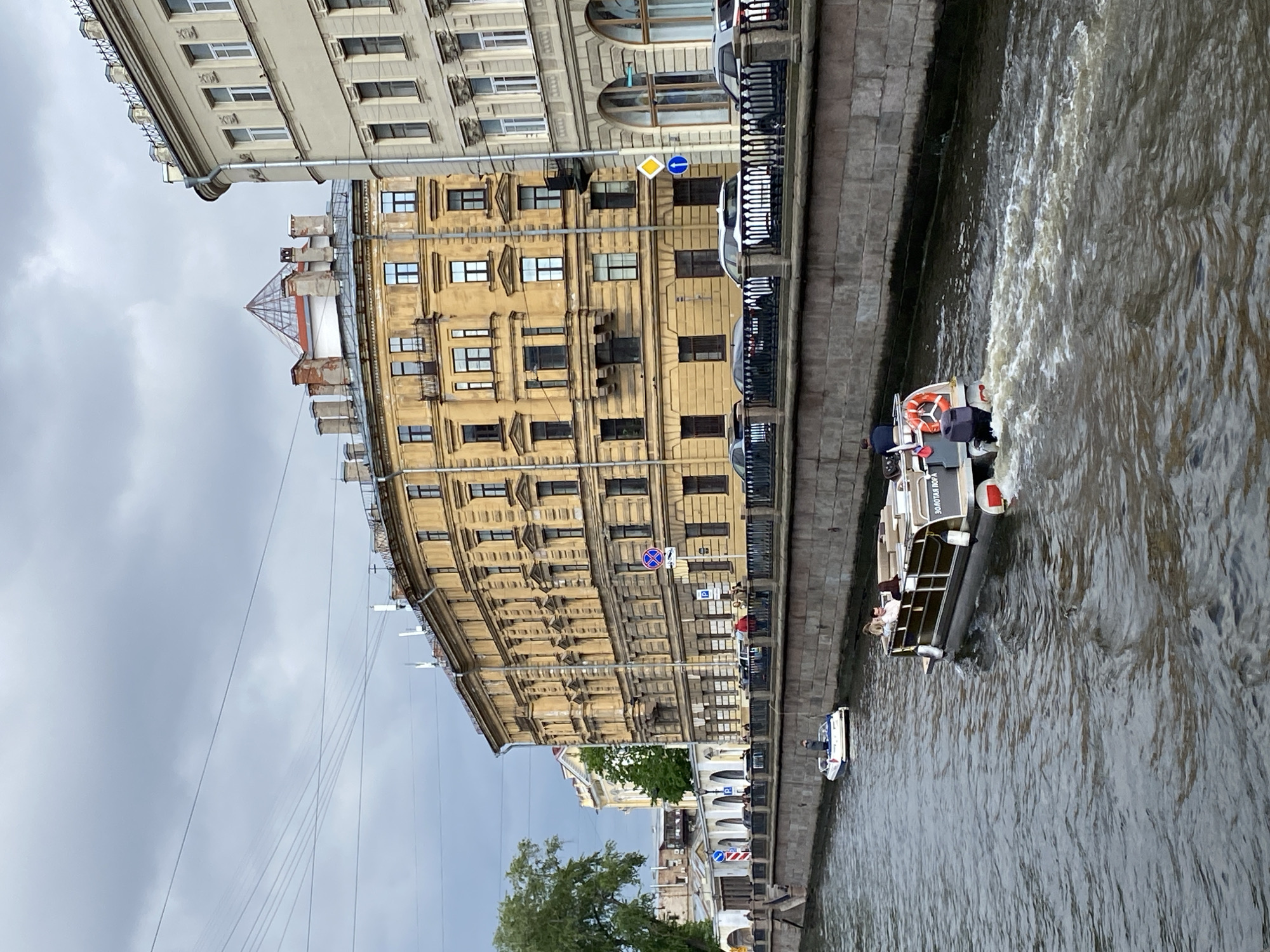 The image shows a canal in Saint Petersburg, Russia. Along the canal, there are several buildings with ornate facades. A sightseeing boat with passengers is moving along the canal. There are cars parked along the canal's edge next to a metal railing. Street signs are visible on the buildings. The sky is overcast. There are trees along the canal.