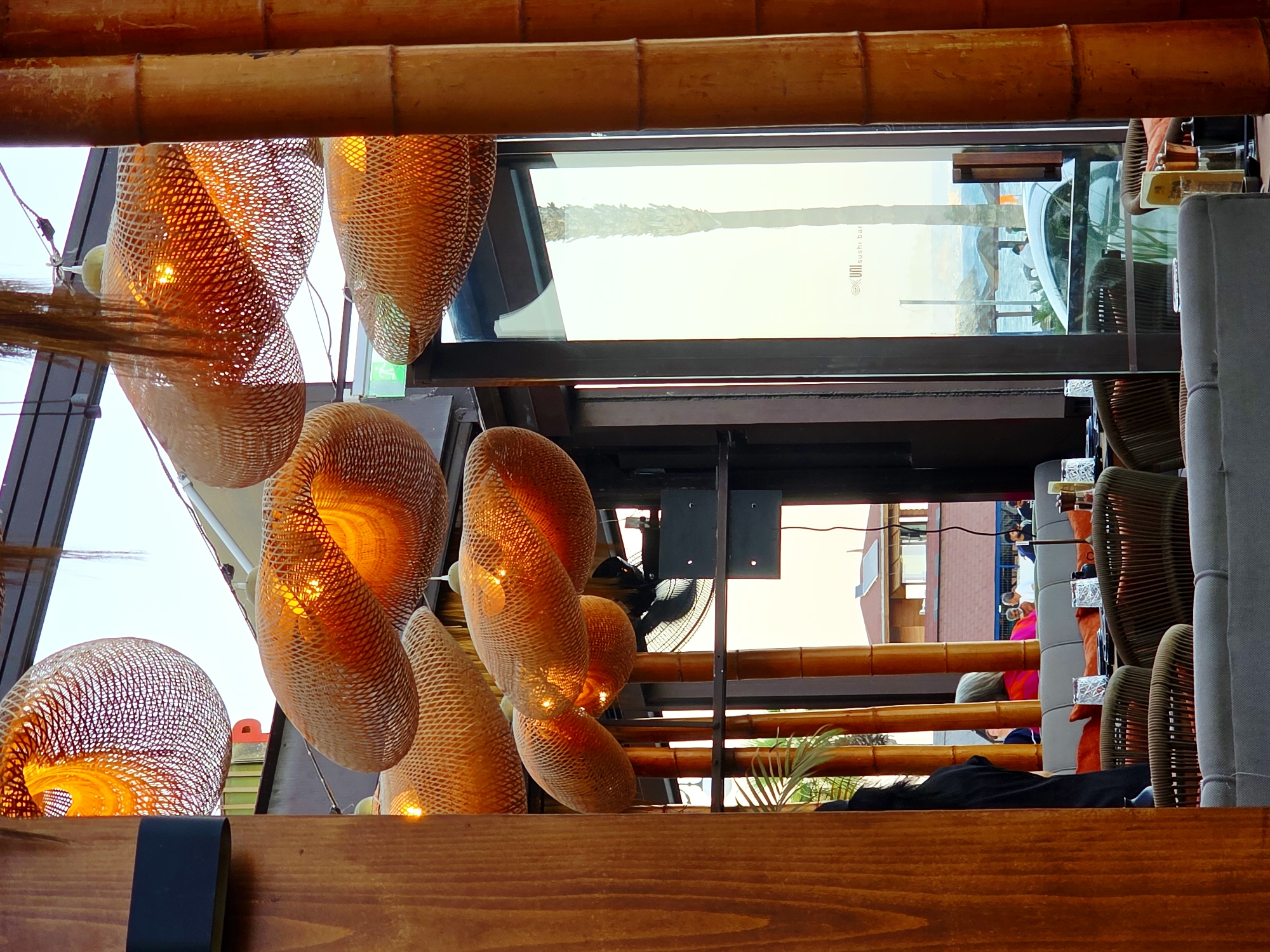 The image depicts the interior of a restaurant with a focus on ambient lighting from multiple woven pendant lamps. These lamps, made of a natural material like bamboo or rattan, cast a warm, orange glow. The restaurant appears to be situated near a waterfront, as suggested by the view through the large windows. In the background, a bright sky, a palm tree, and glimpses of the sea with beach umbrellas are visible. The interior furnishings include comfortable seating with upholstered sofas and woven chairs, suggesting a relaxed dining atmosphere. There are multiple settings for diners, with glasses and tableware arranged on tables.
The scene is likely the "Uni Sushi Bar" in Larnaca, Cyprus. A subtle sign with the logo "Uni Sushi Bar" is visible on the glass window in the middle right of the image. The architectural style with the bamboo elements and the view of the sea strongly aligns with a coastal establishment in Larnaca. People are seated at tables in the background, engaged in dining or conversation, contributing to the lively yet serene ambiance of the restaurant. The overall impression is one of a sophisticated yet comfortable dining experience, enhanced by the unique lighting and proximity to the sea.