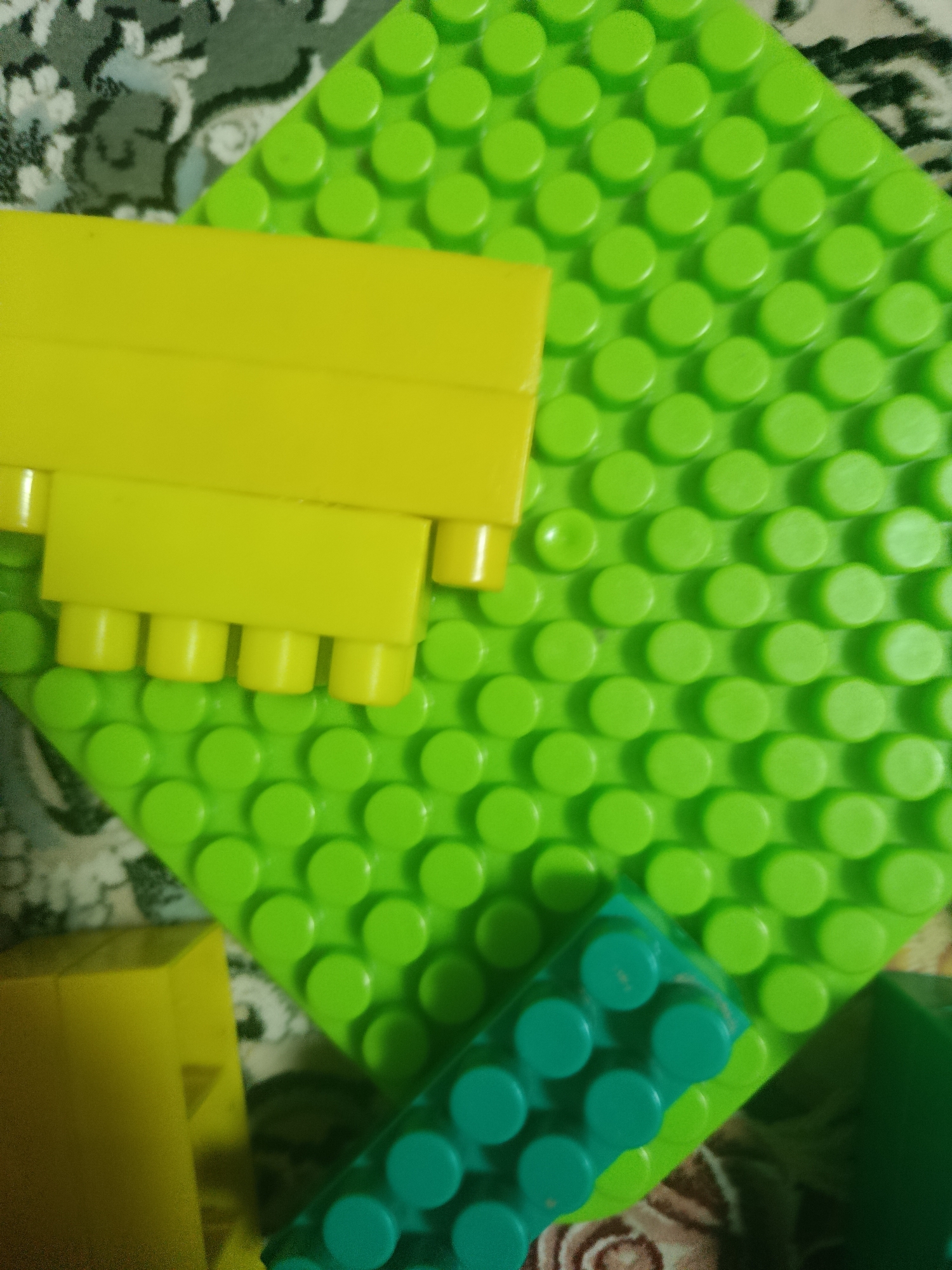 A close-up shot shows several colorful building blocks arranged on a patterned surface. The main subject is a large, bright green building plate with raised circular pegs, a common feature of construction toy sets. On top of this plate, a yellow building block with four cylindrical protrusions is placed, suggesting it's meant to connect to the green base. Another smaller building block, teal in color, is visible in the lower right portion of the image, also featuring pegs for connection. A larger yellow block is partially visible in the bottom left corner. The background consists of a textured fabric with a faded floral or abstract pattern, in shades of green, black, and white. The lighting is natural, casting soft shadows and highlighting the glossy texture of the plastic blocks.