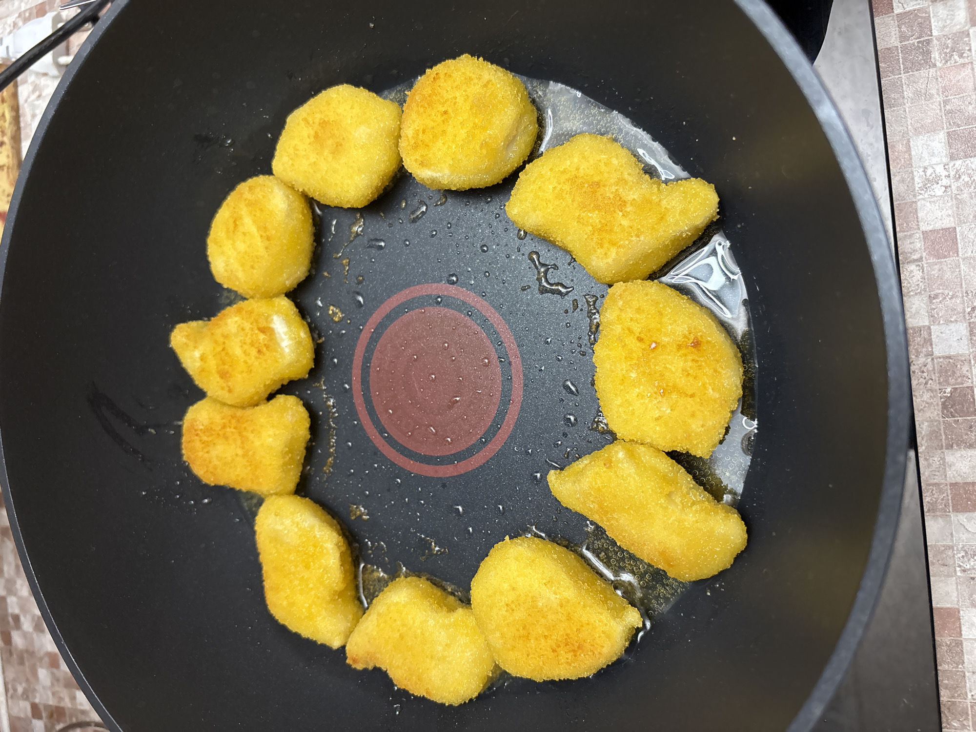 In a black non-stick frying pan, a circular arrangement of golden-brown chicken nuggets is being fried in oil. The nuggets vary slightly in shape and size, some appearing more elongated and others more rounded. They are coated in a crispy breading with visible texture. The oil in the pan is shimmering and shows small bubbles around the nuggets. In the center of the pan is a red and black heat indicator circle. The background shows a countertop with a tiled surface in muted earthy tones. The image appears to have been taken in a domestic kitchen, likely in Ветлужский, Russia, although there are no specific indicators of this location.