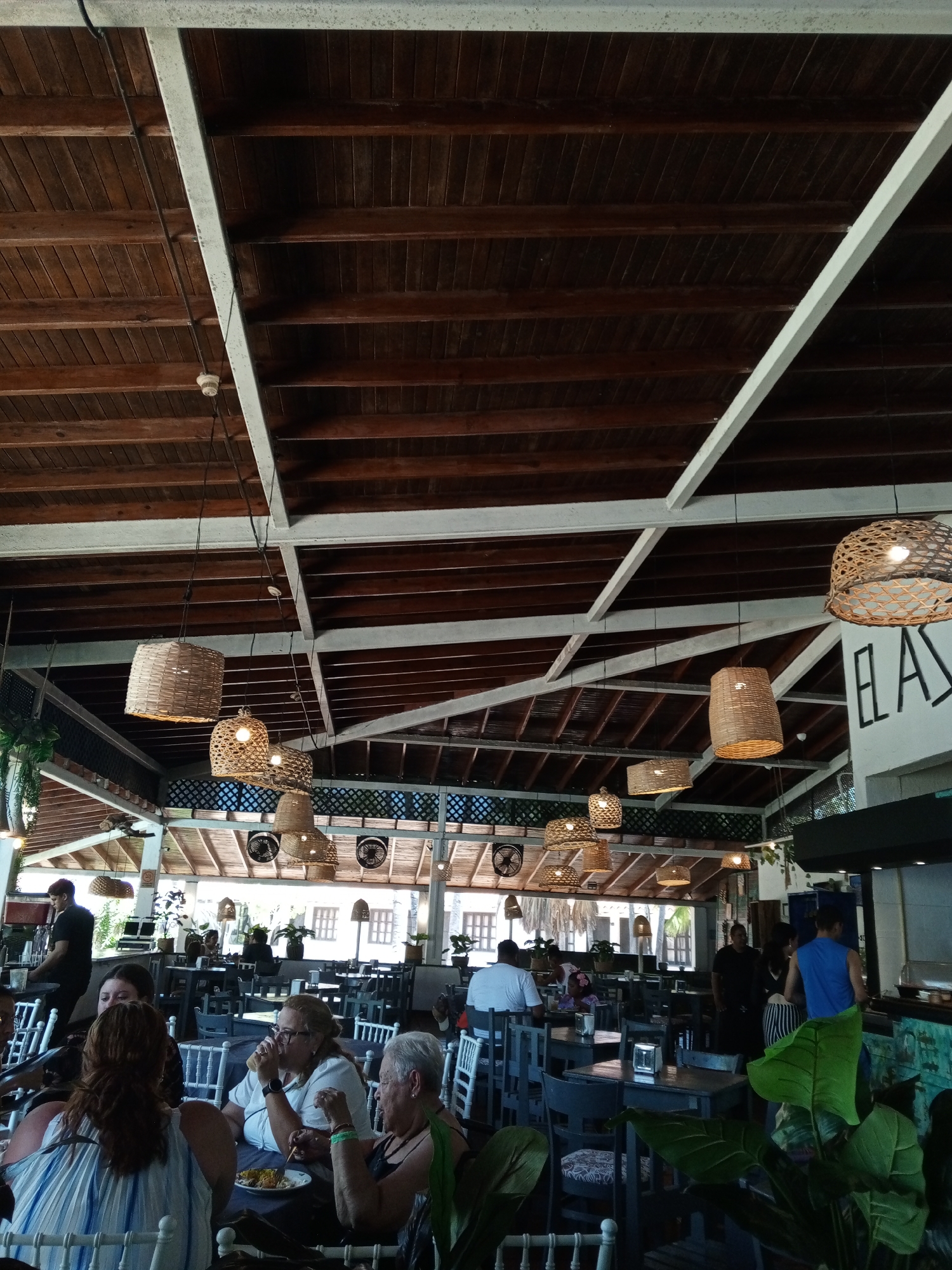 The image depicts the interior of a restaurant, potentially located in Porlamar, Venezuela, given the request. The space is characterized by a high ceiling with exposed wooden beams and white structural supports. Woven lamps hang at varying heights throughout the restaurant, illuminating the tables below. Numerous patrons are seated at tables, some appearing to be engaged in conversation and enjoying meals. Waitstaff can be seen moving through the space, attending to the customers. The decor includes potted plants and a color palette featuring white, wood tones, and neutral colors. The overall ambiance appears relaxed and informal. The image captures a scene of a bustling restaurant with customers enjoying the ambiance and services.