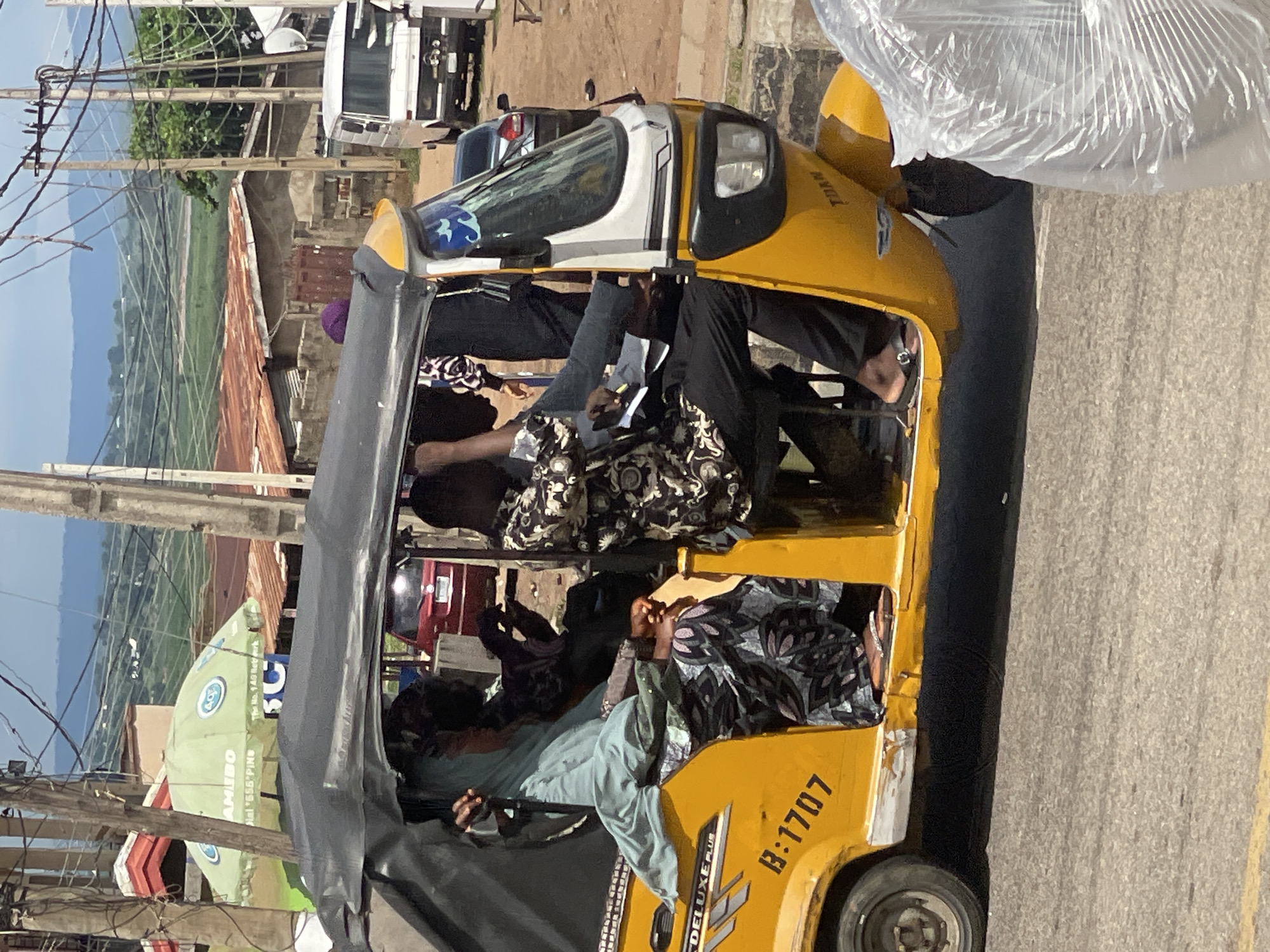 A yellow three-wheeled auto-rickshaw, identified by the license plate B:1707 and the text "DELUXE PLUS," is driving on a paved road. The auto-rickshaw has a black canopy and its doors are open. Inside, a woman wearing a light blue headscarf and a patterned dress is seated on the left. To her right, a man in a patterned shirt and black pants is seated, holding a pen and paper. Another person, partially visible, is seated behind the driver. In the background, there are various buildings with corrugated metal roofs, utility poles with numerous wires, and lush green hills in the distance under a bright, partly cloudy sky. A white van is parked on the right side of the road.