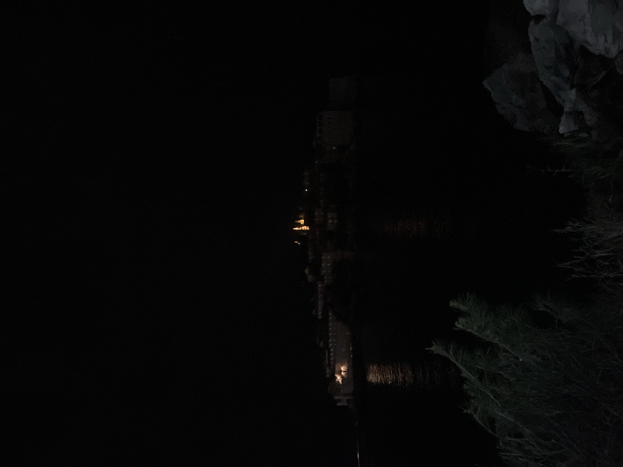 The image, taken at night, shows a dark body of water, possibly a bay or lake, with reflections of lights in the water. A building with multiple floors is dimly lit and reflected in the water. The right side of the frame shows rocky terrain and vegetation. The scene appears to be in or near a built environment, potentially along a coastline. The location, based on the prompt, is Rađenovići, Montenegro.