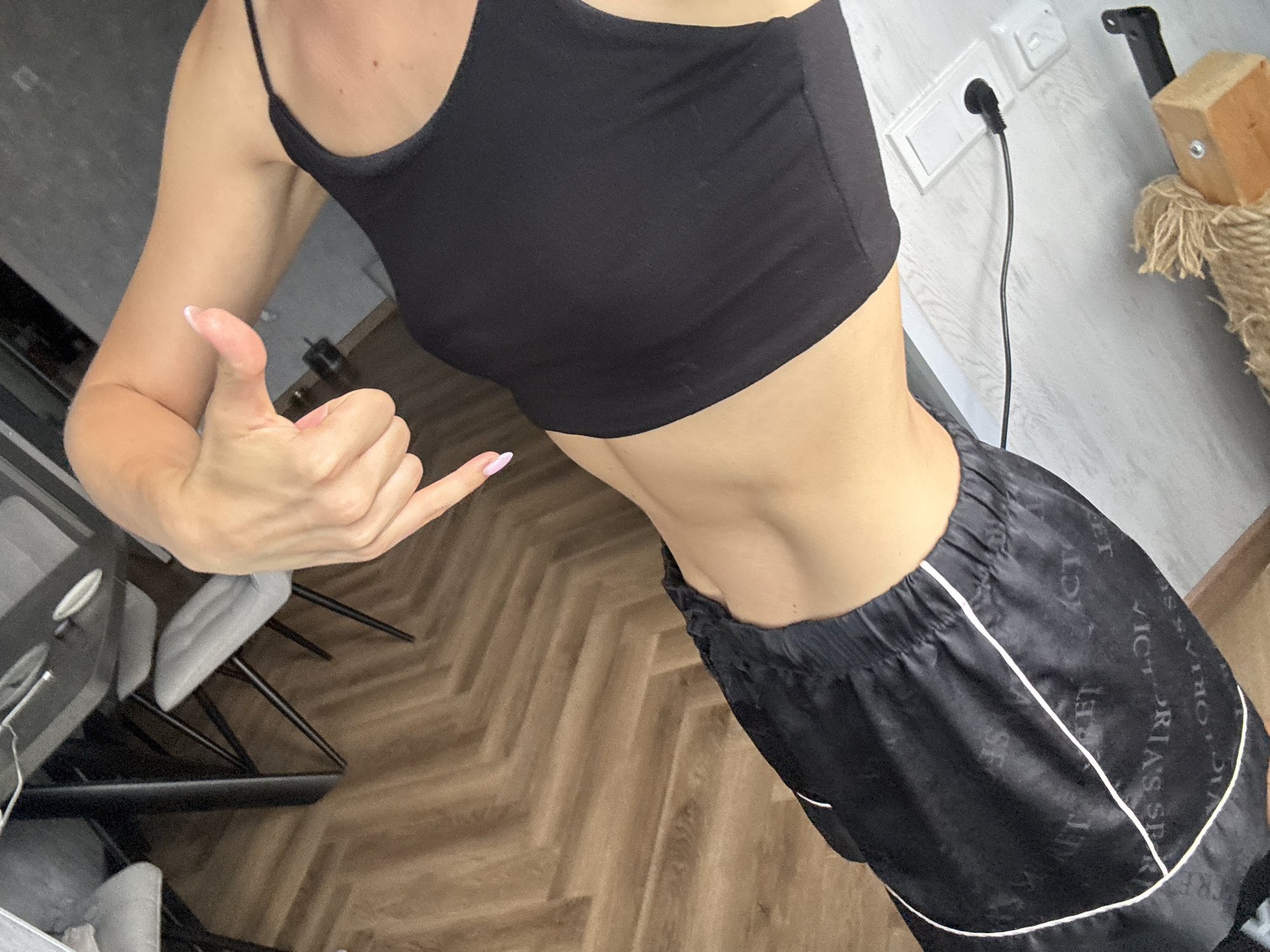A person is captured from the torso down, showing their midsection and arms. They are wearing a black tank top and black pajama shorts with white trim. The shorts have a repeating pattern of the words "VICTORIA'S SECRET". The person is standing on a wooden floor with a herringbone pattern. In the background, there is a wall with electrical outlets and a plug. To the left, part of a dining table and chairs can be seen. The lighting suggests it is daytime. The person is giving a "shaka" hand gesture with their left hand, with their thumb and pinky finger extended. Their right hand is resting on their hip. The overall mood is casual and relaxed.