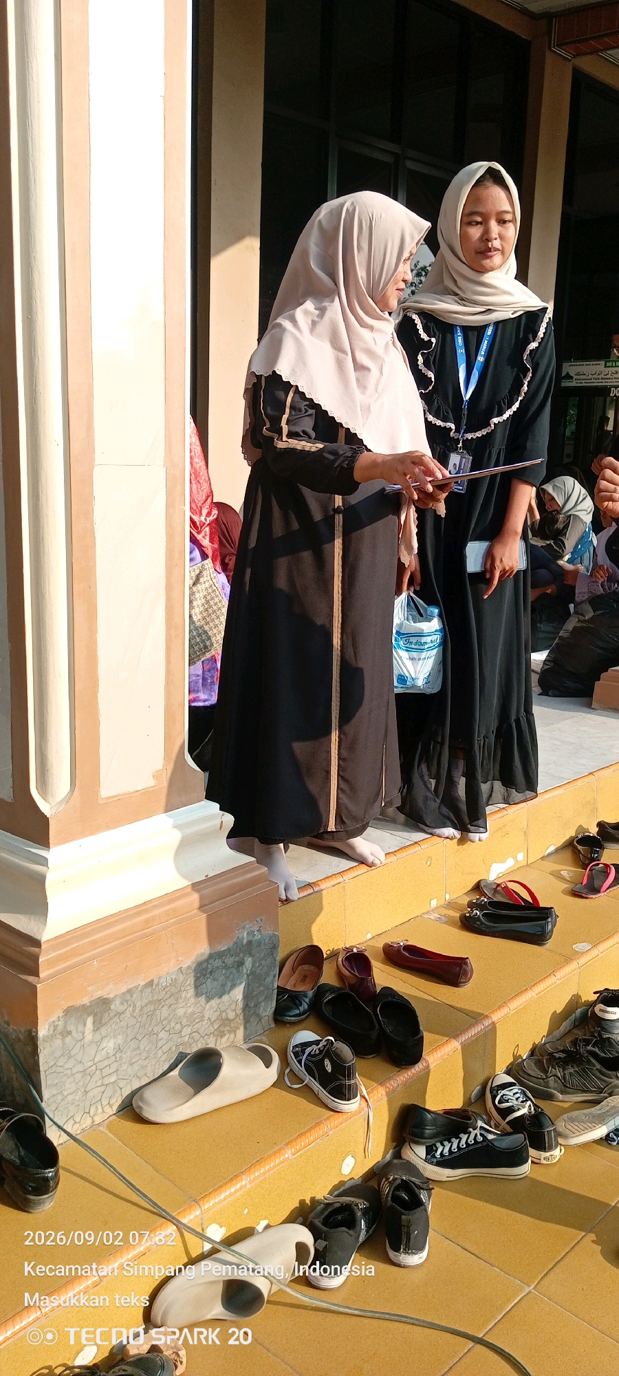 The image shows two young women wearing hijabs and long, dark dresses, standing on the steps of a building in Simpang Pematang, Indonesia. They are facing away from the camera, with one woman on the left holding a piece of paper and the other on the right holding a phone. Several pairs of shoes are scattered on the steps, including sandals, flats, and sneakers. The lighting suggests it is daytime, with shadows cast by the building's pillars. A plastic bag with an "Indomaret" logo is visible near the woman on the left. The building appears to be a place of worship or public gathering, given the presence of footwear outside. A watermark at the bottom of the image indicates the date as "2026/09/02 07:32" and the location as "Kecamatan Simpang Pematang, Indonesia," along with "TECNO SPARK 20."