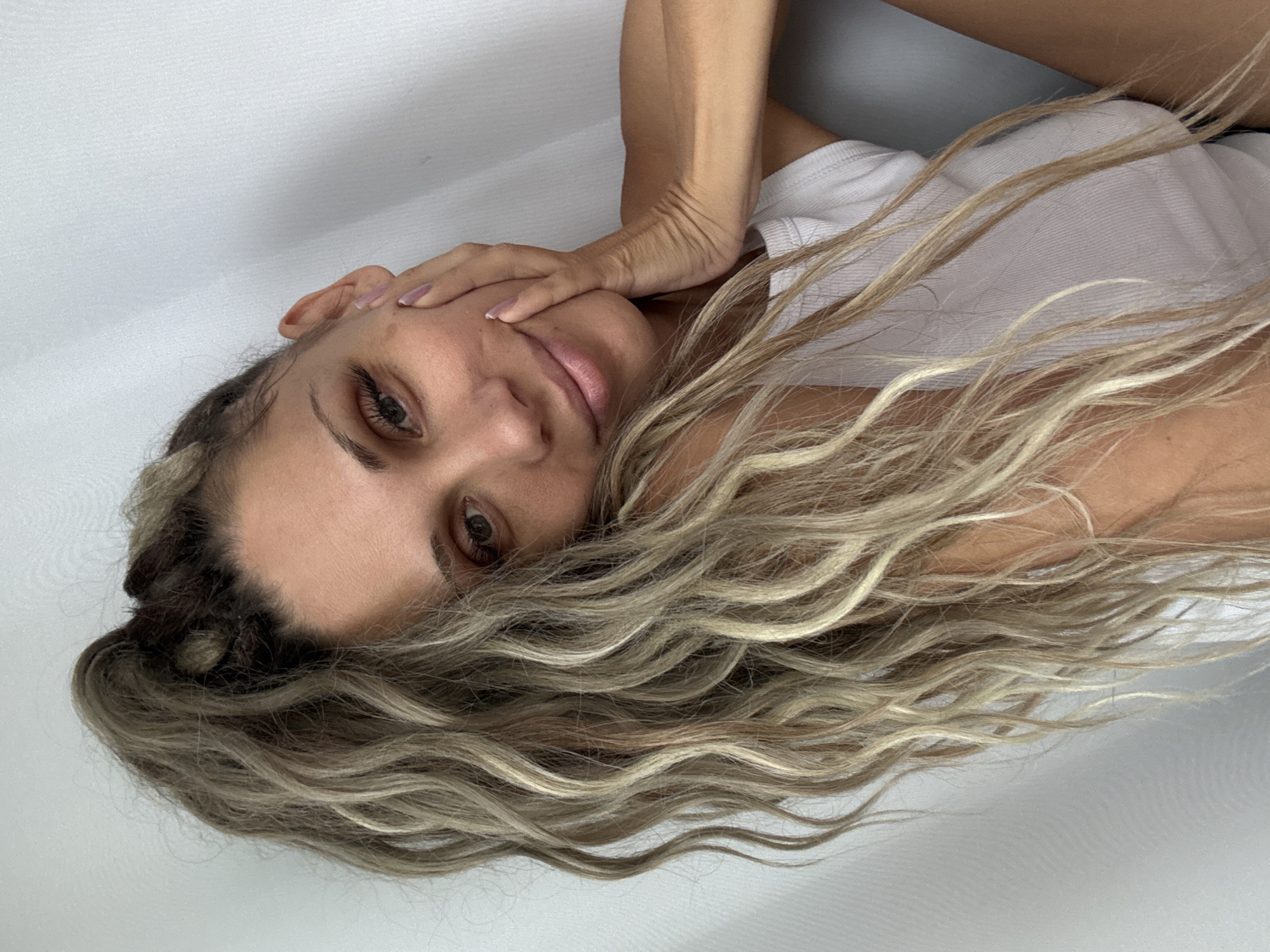 A woman with long, wavy, blonde and brown hair in a high ponytail is shown in a close-up, medium shot from the chest up. She is resting her right cheek on her right hand, which is placed on her face, and she is wearing a white tank top. The background is a plain, light gray wall. The image does not contain any elements that indicate the location in Rusanovo, Russia.