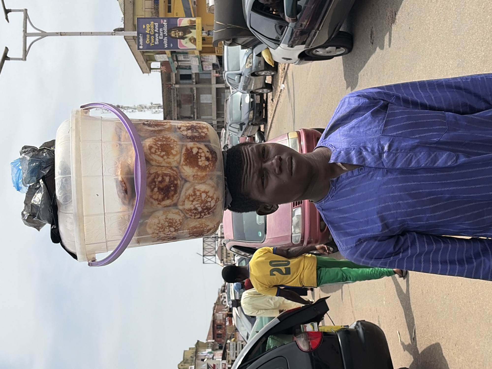 A young man wearing a blue striped tunic and carrying a bucket of pastries on his head stands on a dusty road in Jos, Nigeria. He is looking towards the camera, his expression neutral. The bucket, made of clear plastic with a purple rim, is filled with small, round pastries that appear to be puffed and golden brown. A black plastic bag is tucked into the top of the bucket. In the background, several cars are visible, indicating a street scene with moderate traffic. A billboard for "Jollof+" is also present on the right side of the image. The overall scene suggests a bustling urban environment.