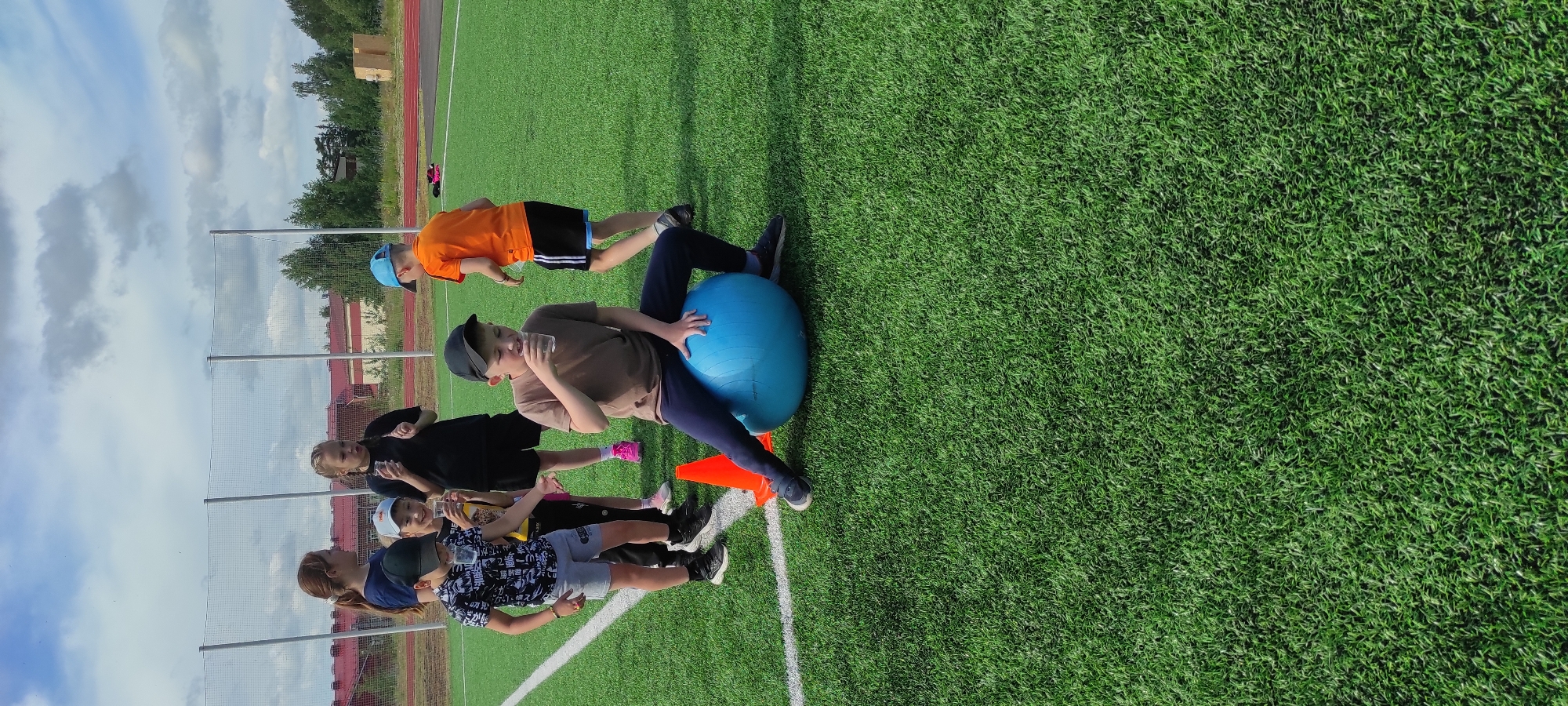 In Noviy, Russia, a group of children and a young woman are gathered on a green artificial turf sports field. A boy, wearing a brown t-shirt and dark pants, is sitting on a large blue exercise ball, drinking from a clear plastic cup. He is wearing a black baseball cap. Beside him, an orange cone is visible. To the left of the boy on the ball, another boy in a black and white patterned t-shirt and grey shorts is also holding a clear cup. Behind them, a young woman with red hair pulled back in a ponytail stands near a girl with braided hair, who is also holding a cup. Other children are also present, some holding cups. The background shows a net fence, some trees, and buildings. The sky is partly cloudy.