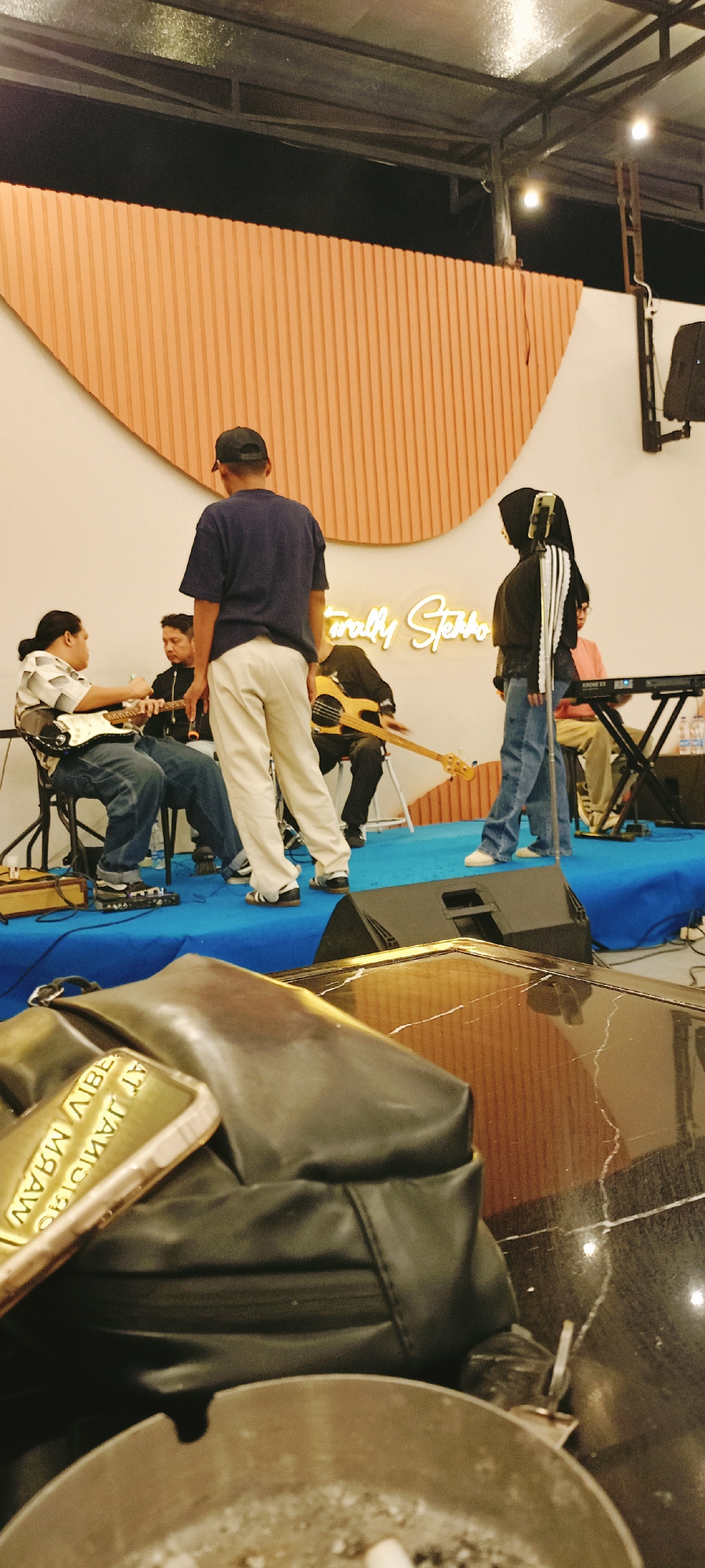 The image shows a band performing on a small stage, likely at a venue in Lhokseumawe, Indonesia, given the prompt. The stage is covered with a blue cloth. From left to right, a man sits playing an electric guitar, another man sits holding a microphone, a man with his back to the camera stands wearing a dark blue shirt and white pants, a man plays a bass guitar, and a woman in a headscarf stands next to a man playing a keyboard. Behind the band is an orange wavy backdrop and a neon sign reading "Naturally Sketch". A speaker sits at the front of the stage. The foreground shows a black leather bag on a table. The overall lighting suggests an indoor performance space.