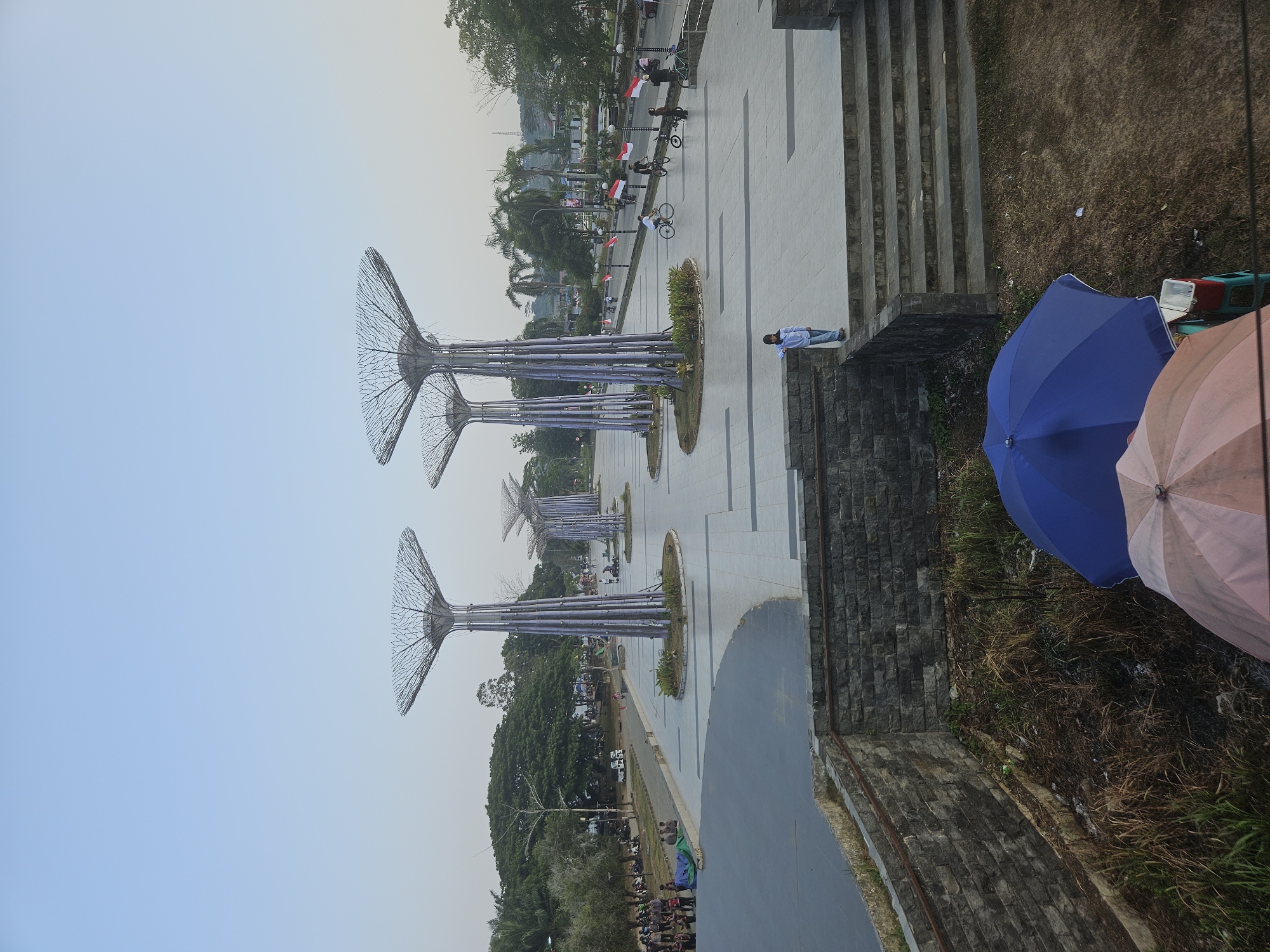 The image depicts an outdoor public space in Teluk Dalam, Indonesia, featuring several large, tree-like sculptures with intricate metal canopies. These sculptures are arranged along a paved promenade lined with small Indonesian flags. In the background, there are trees, buildings, and a road with some traffic. People are visible throughout the scene, including individuals walking, cycling, and gathered in groups. In the foreground, a stone wall with a rough texture borders an area with dry grass and foliage. Two umbrellas, one blue and one pink, are visible in the lower right corner, along with a cooler box on a stool. The sky is clear and pale blue, suggesting either early morning or late afternoon.