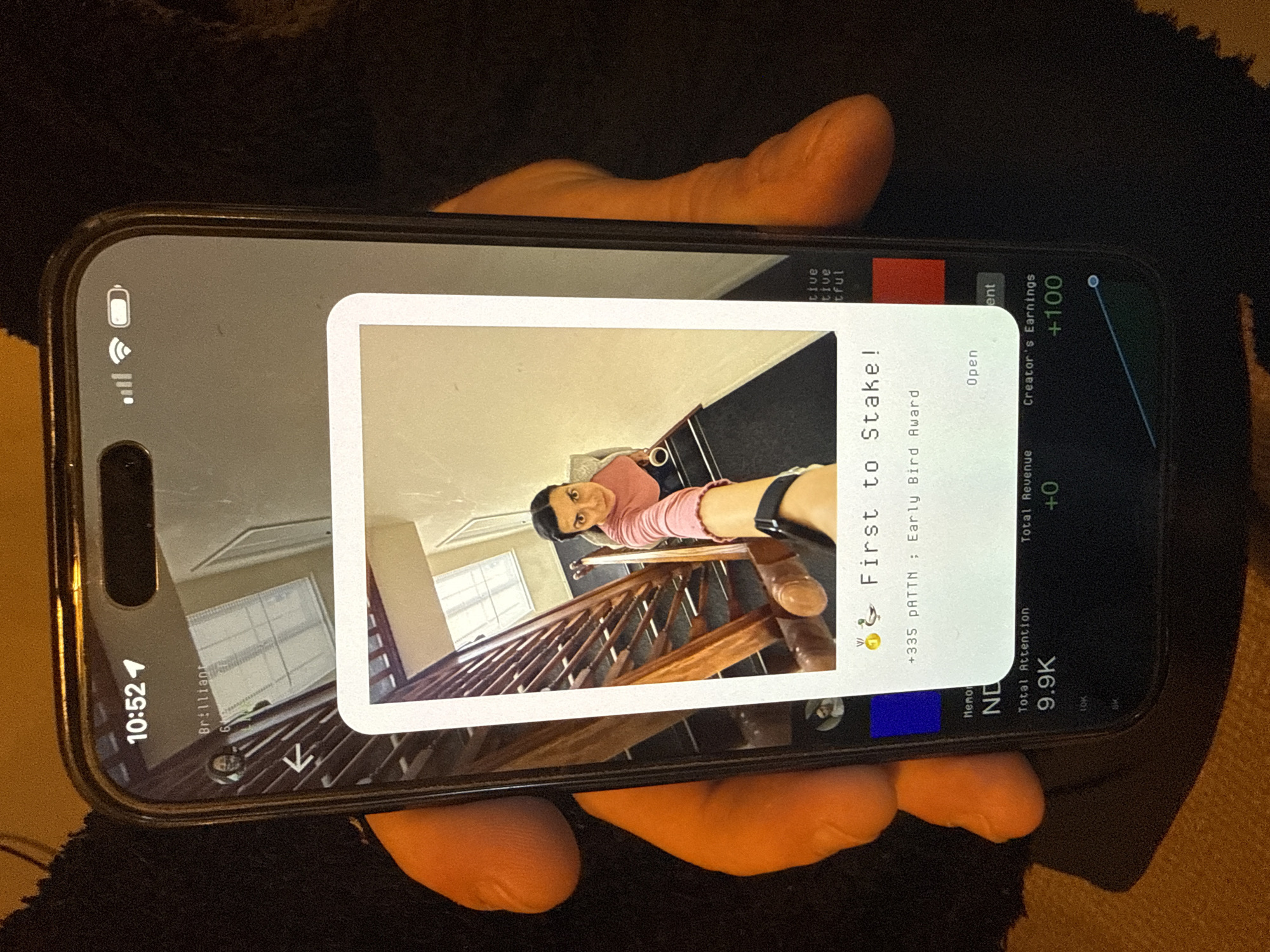 A hand is holding a smartphone displaying a social media post. The post features a woman on a staircase and is titled "First to Stake! Early Bird Award." The phone's screen also shows metrics such as "Total Attention: 9.9K," "Total Revenue: +0," and "Creator's Earnings: +100." The phone displays the time as 10:52. The location is not indicated on the screen or image.