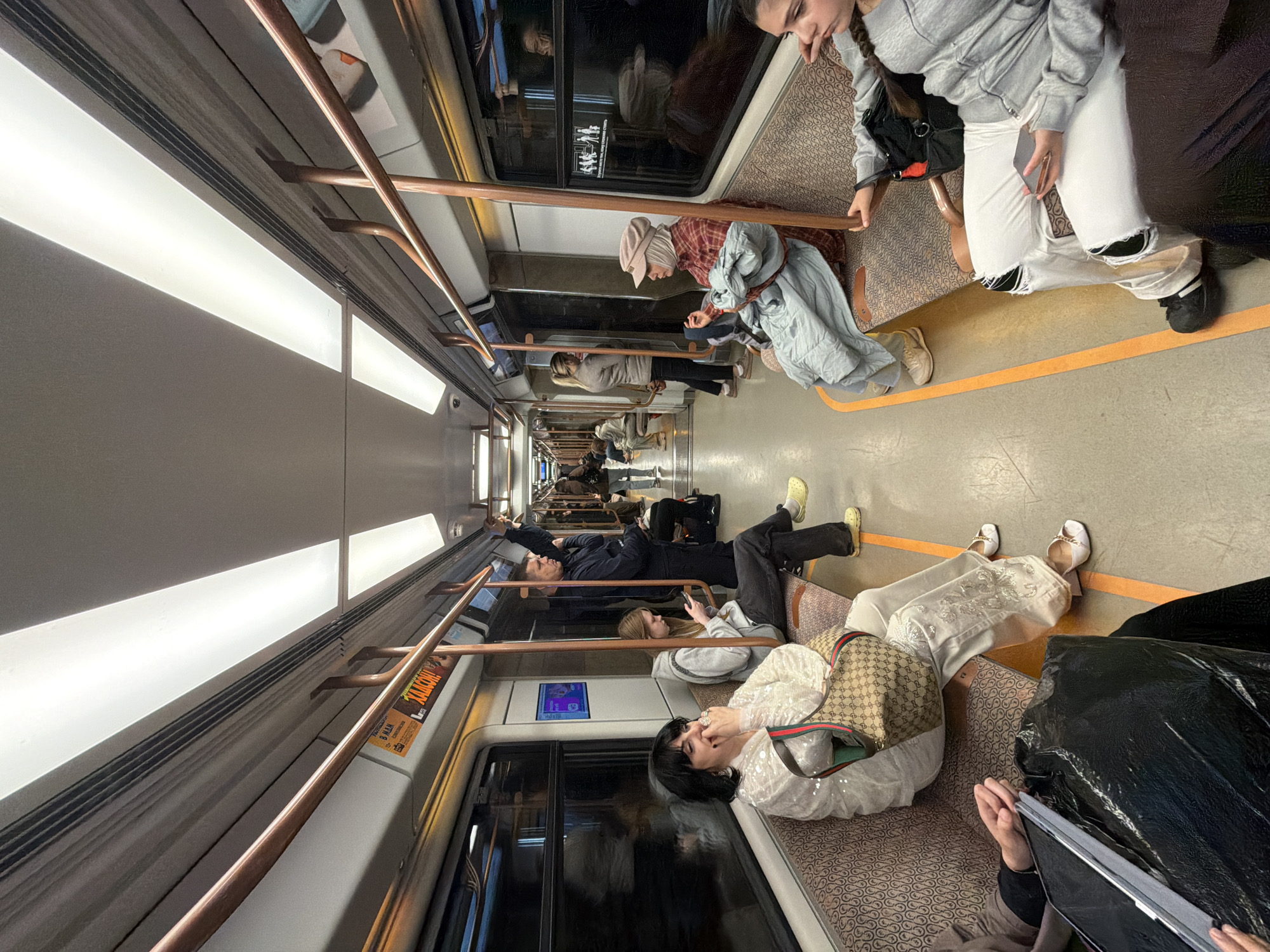 The image depicts the interior of a subway car in Moscow, Russia. Several passengers are seated along the length of the car. They appear to be engaged in individual activities such as looking at their phones, sleeping, or talking on a laptop. The subway car features patterned seating, copper handrails, and linear fluorescent lighting. The overall ambiance suggests a typical urban commute.