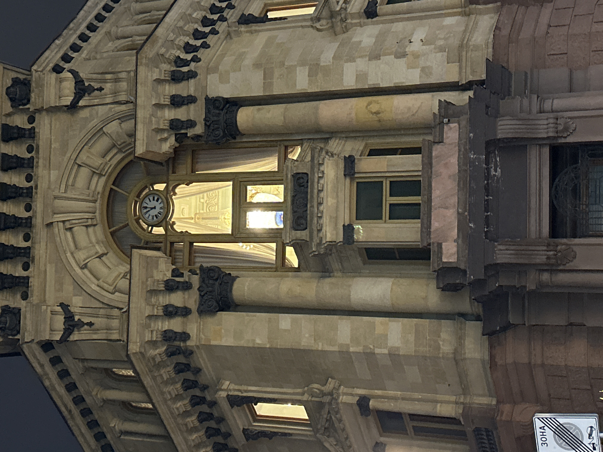A medium shot captures a corner of an ornate, classical-style building at night. The building is constructed of light-colored stone blocks, with darker, intricate decorative elements along the roofline and around windows. A large, arched window dominates the central section, featuring heavy, golden curtains pulled back to reveal illuminated interior spaces. Above this, a circular clock with Roman numerals is visible, its hands pointing to approximately 11:52. Flanking the central window are tall, classical columns with decorative capitals.
On the left side of the image, a smaller rectangular window is lit from within. Below the main arched window, there is a bay window structure with multiple panes. The lower portion of the building visible in the frame features darker stone and a more imposing entrance with a decorative cornice. A street sign with Cyrillic text and a symbol indicating a no-stopping zone is positioned in the lower left corner. The sky above is dark and indistinct, indicating nighttime.