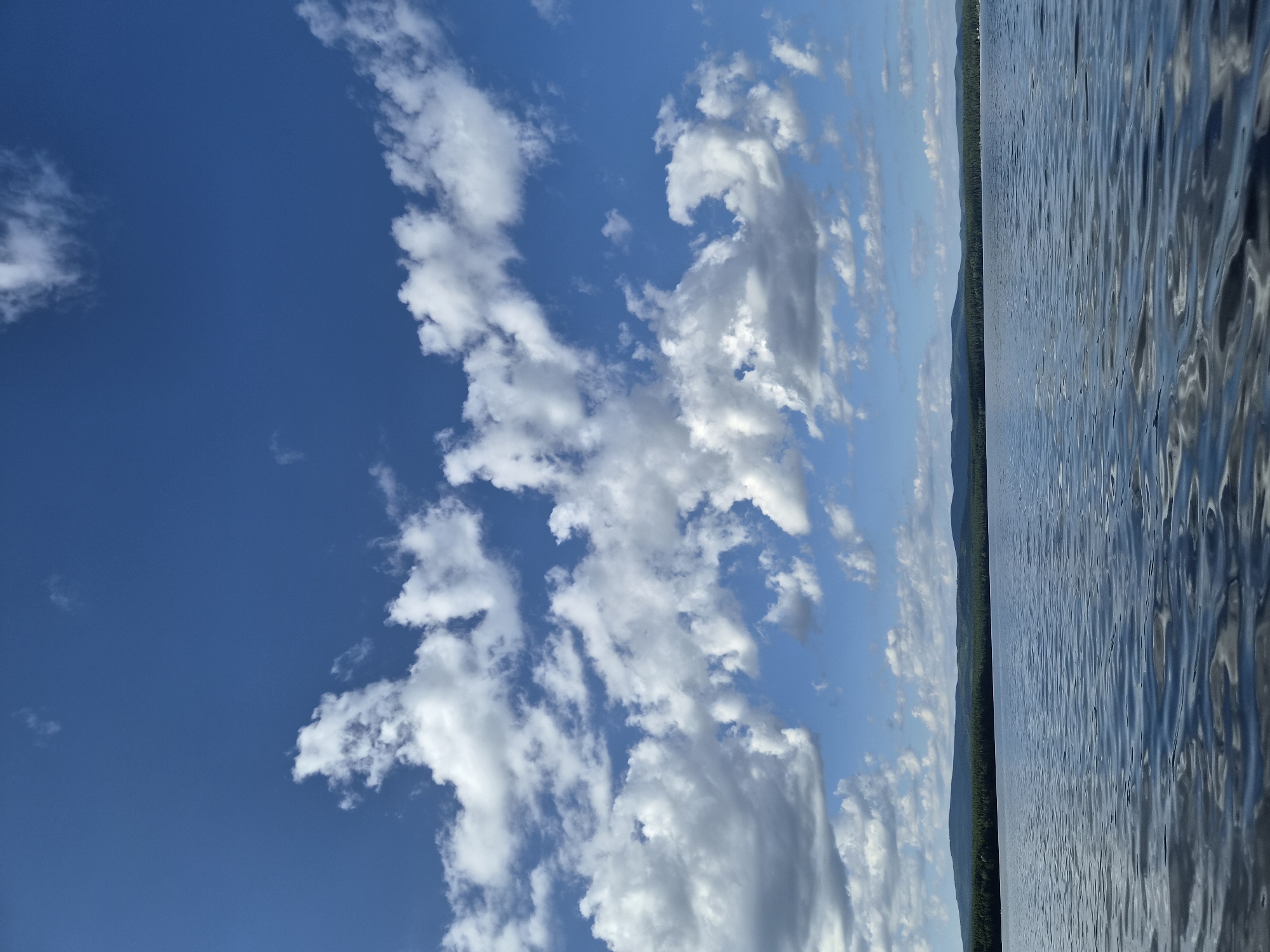 The image depicts a wide expanse of calm water, likely a lake, under a bright blue sky dotted with fluffy white clouds. In the distance, a dense line of dark green forest stretches across the horizon, with rolling hills or mountains visible behind it. The water's surface is gently rippled, reflecting the sky and clouds. The scene appears to be set in the city of Areyskiy, Russia, evoking a sense of natural tranquility. There are no people or animals visible in the image.