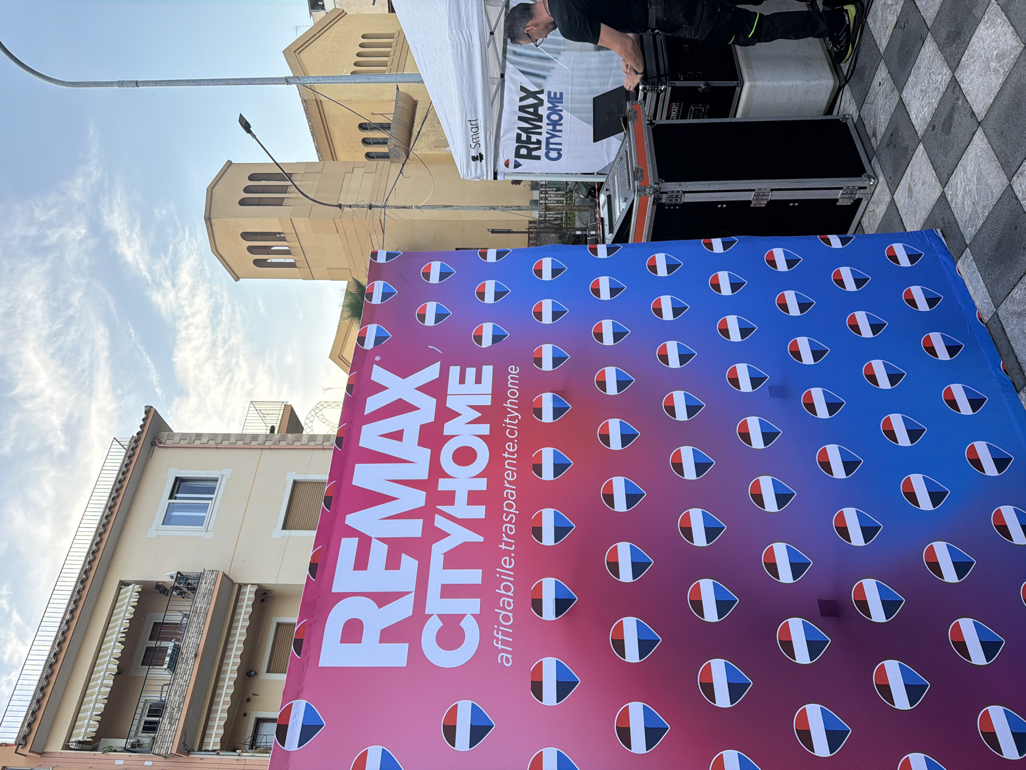 The image shows a marketing display for "REMAX CITYHOME" in Chianchitta, Italy. A large banner with a gradient color scheme (pink to blue) and repeated teardrop-shaped design stands in front of a building and a street. A man is partially visible, standing under a white tent branded with the "REMAX CITYHOME" logo, operating what seems to be audio equipment. The background features buildings with architectural details and a partly cloudy sky. The ground where the banner and tent are set up is paved with a checkerboard pattern. The additional text "affidabile.trasparente.cityhome" is also visible on the banner.