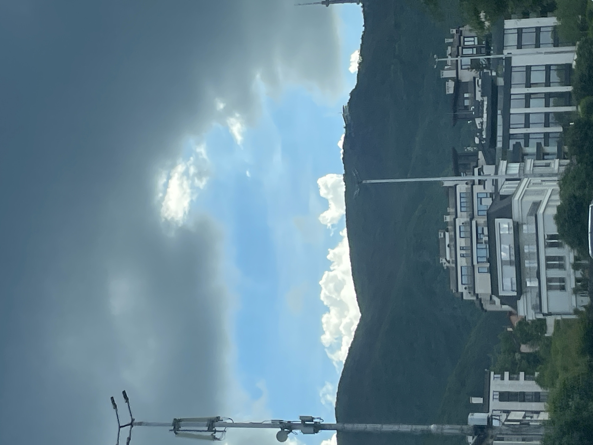 The image shows a view of buildings and vegetation situated on a hillside. A mountain or large hill is visible, partially obscured by clouds in the sky. The buildings appear to be modern and well-maintained. In the foreground, there is some sort of equipment on a pole. The image is rotated 90 degrees counterclockwise.