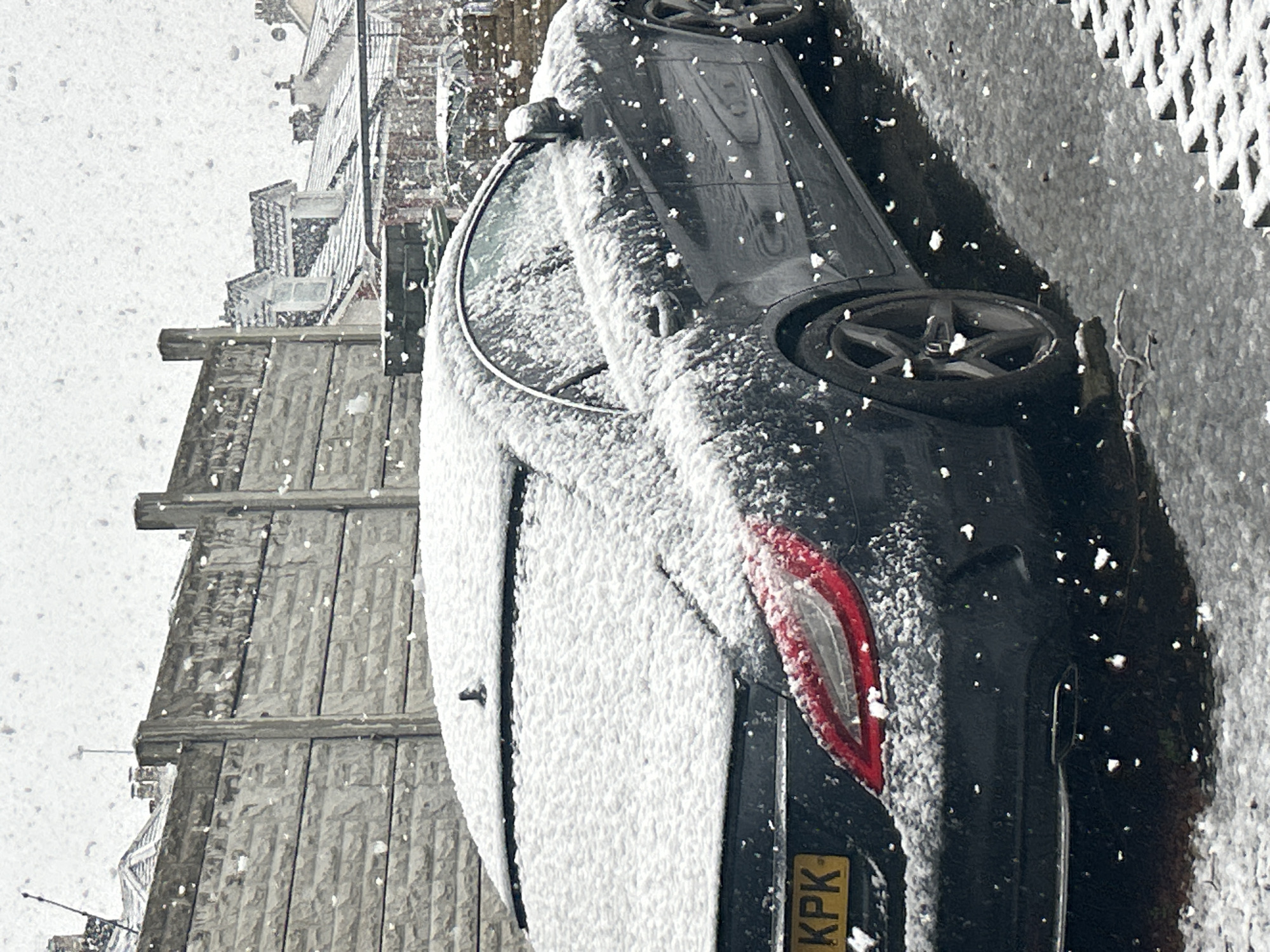 A black car is covered in snow. The car is parked on the side of the road next to a stone wall. Snow is falling heavily. The license plate of the car is visible, and reads "KPK". A residential building can be seen in the background. The location is Glasgow, United Kingdom.