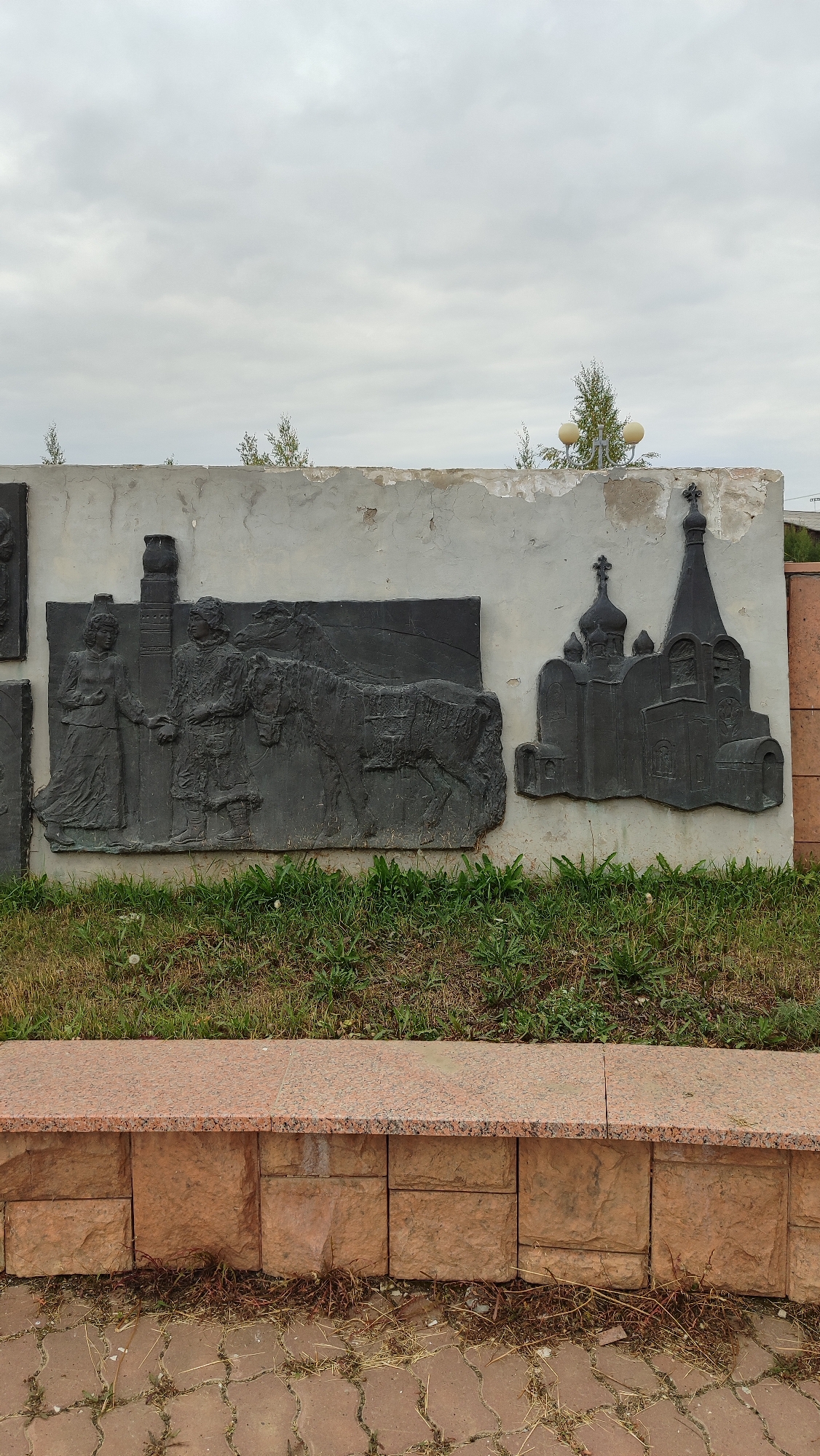 The image shows a bas-relief sculpture on a weathered concrete wall in Mirny, Russia. The sculpture depicts two figures, one a woman in a long dress, and the other a person in traditional clothing, standing next to a horse. To the right of this scene is a depiction of a church with multiple domes and a cross. The wall is mounted on a stone base, with a granite bench in front of it. Green grass grows around the base of the wall, and the ground in front of the bench is paved with interlocking stones. The sky above is overcast with clouds.