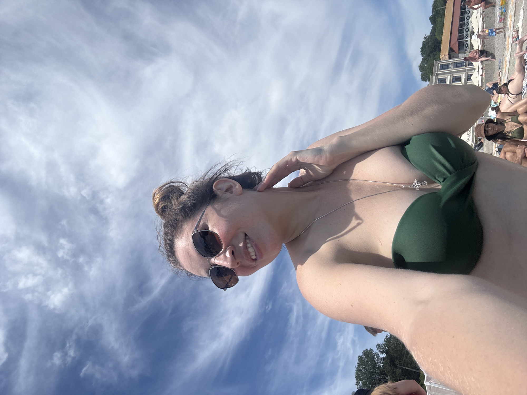 A woman is taking a selfie on a beach under a cloudy sky. She is wearing a green bikini top and sunglasses. Her hand is touching her neck. In the background, there are other people on the beach, umbrellas, buildings, and trees. The scene appears to be a resort area. The location is identified as Odesa, Ukraine.