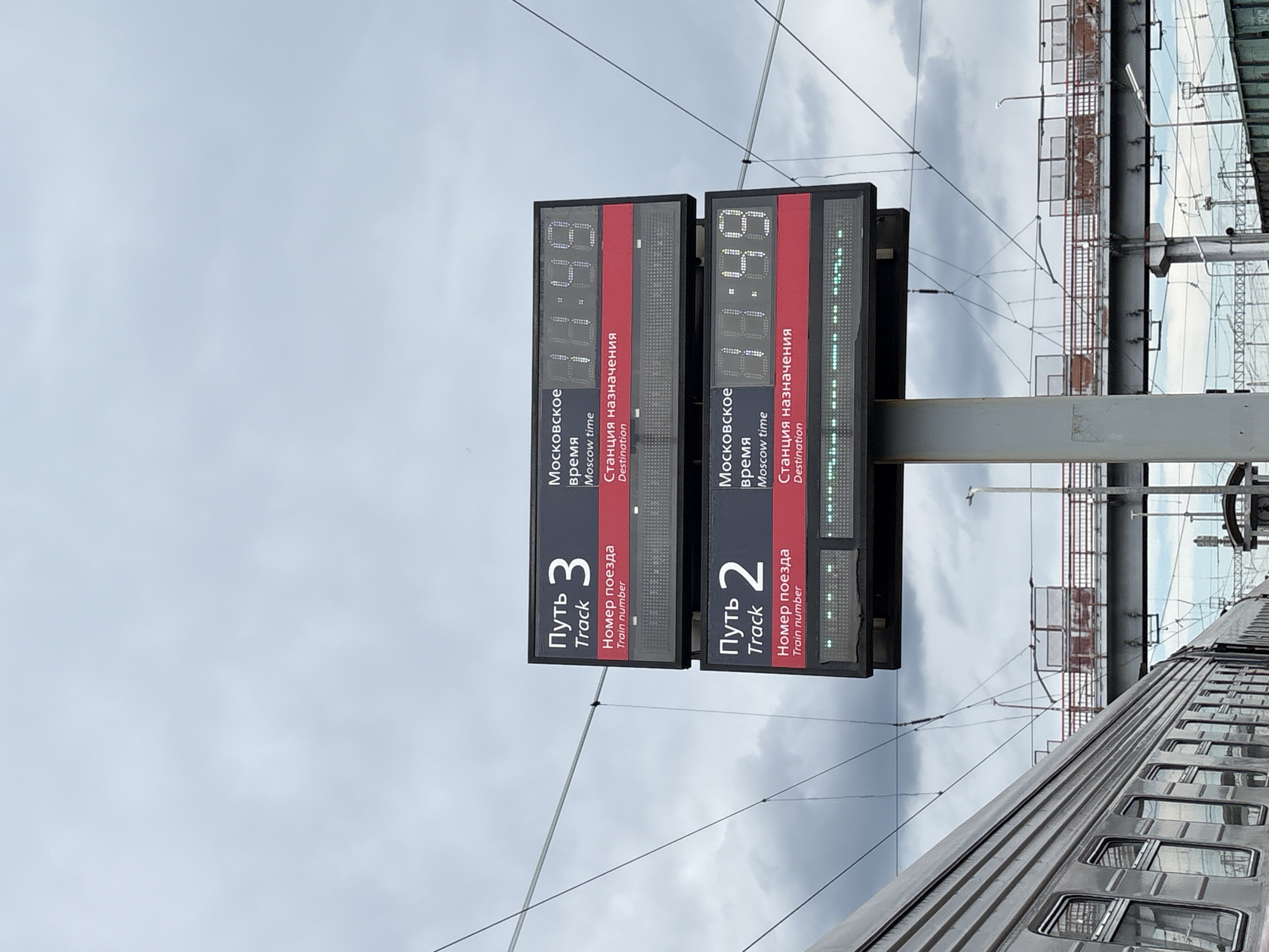 The image shows two digital display boards at a train station, likely in Povorino, Russia, under a cloudy sky. The boards display train information, including track numbers, train numbers, Moscow time, and destination stations. The top board shows "Track 3" and the bottom board shows "Track 2". Both boards display the Moscow time as "11:49". The destination display for both tracks is currently blank, indicated by a line of green lights. A portion of a grey train is visible on the left side of the image, running parallel to the display boards. In the background, a pedestrian bridge with railings and overhead electrical wires are visible, suggesting an active railway environment. The overall scene is one of a functional train station.
