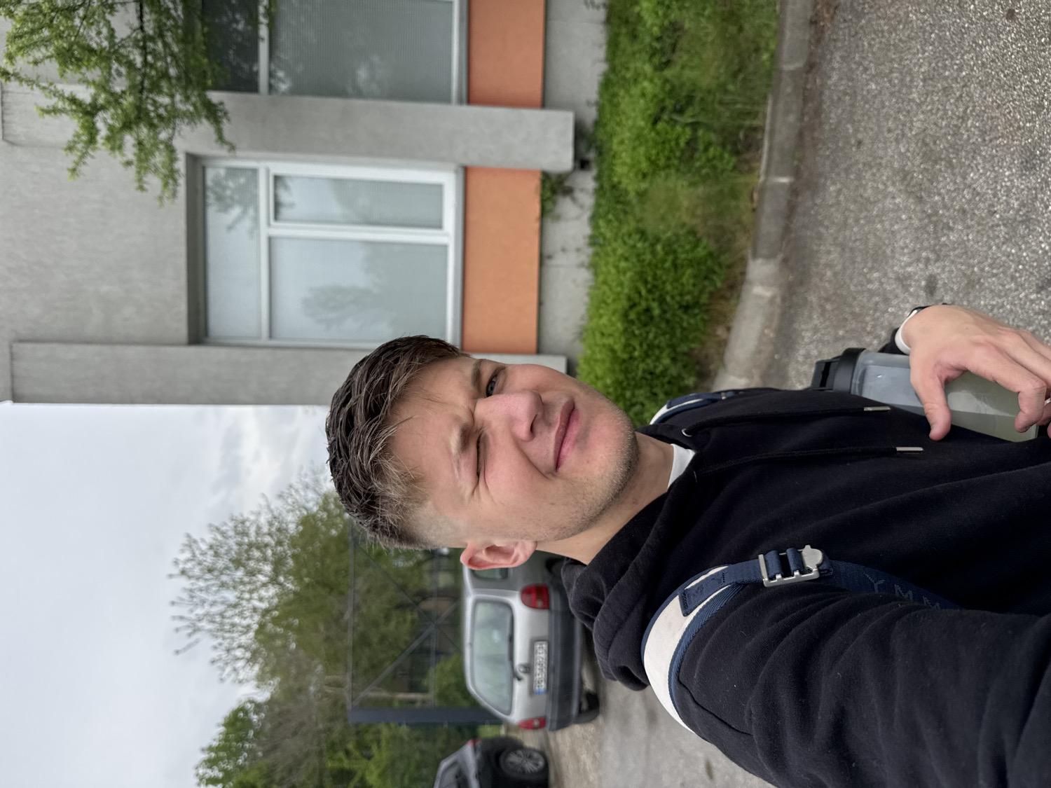 A young man is captured in a selfie, with the image rotated 90 degrees clockwise. He faces the camera, winking with his right eye and exhibiting a casual, slight smirk. He has short, neatly styled dark hair, light skin, and light stubble. Dressed in a black hoodie over a white t-shirt, he has a blue and white strap, possibly from a backpack, visible over his right shoulder. In his right hand, he holds a translucent bottle with a black lid, containing a light yellow-green liquid. A silver ring is visible on his left ring finger.
The background depicts an urban or suburban outdoor setting under an overcast, grey sky. To the man's left, a multi-story building features a facade with alternating grey and orange sections and multiple windows with white frames. Green tree foliage is visible near the top left of the building. To his right, an asphalt road or parking area stretches back, lined with green grass and shrubs. Several cars are parked in the distance, including a silver car with a partially visible European-style license plate, confirming the location as consistent with Plovdiv, Bulgaria. The overall impression is a candid, daytime shot on a cloudy day.