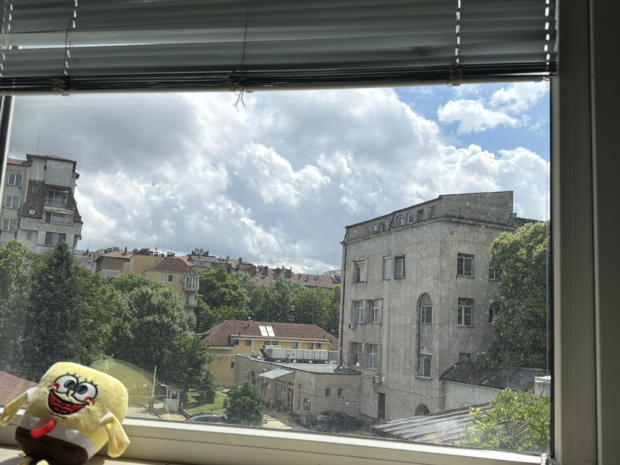 The image presents a daytime view from a window overlooking a cityscape in Sofia, Bulgaria. Horizontal window blinds are partially drawn at the top. On the window sill in the lower-left corner sits a plush Spongebob Squarepants toy, smiling.
The outdoor scene is dominated by a partly cloudy sky with large white cumulus clouds, indicating fair weather. Below, an urban landscape unfolds, characterized by a mix of buildings and lush green trees. To the left, a tall, multi-story concrete building with numerous windows and balconies is visible. On the right, a prominent, light-colored stone building with distinct arched windows on its lower floors stands. Beneath this building, a flatter structure with air conditioning units on its roof and a dark car parked nearby can be seen. In the background, more residential buildings with red-tiled roofs are interspersed with dense tree cover, contributing to a layered urban panorama. A small guard booth is also discernible among the trees in the middle distance. No specific activity or visible text is depicted in the image.