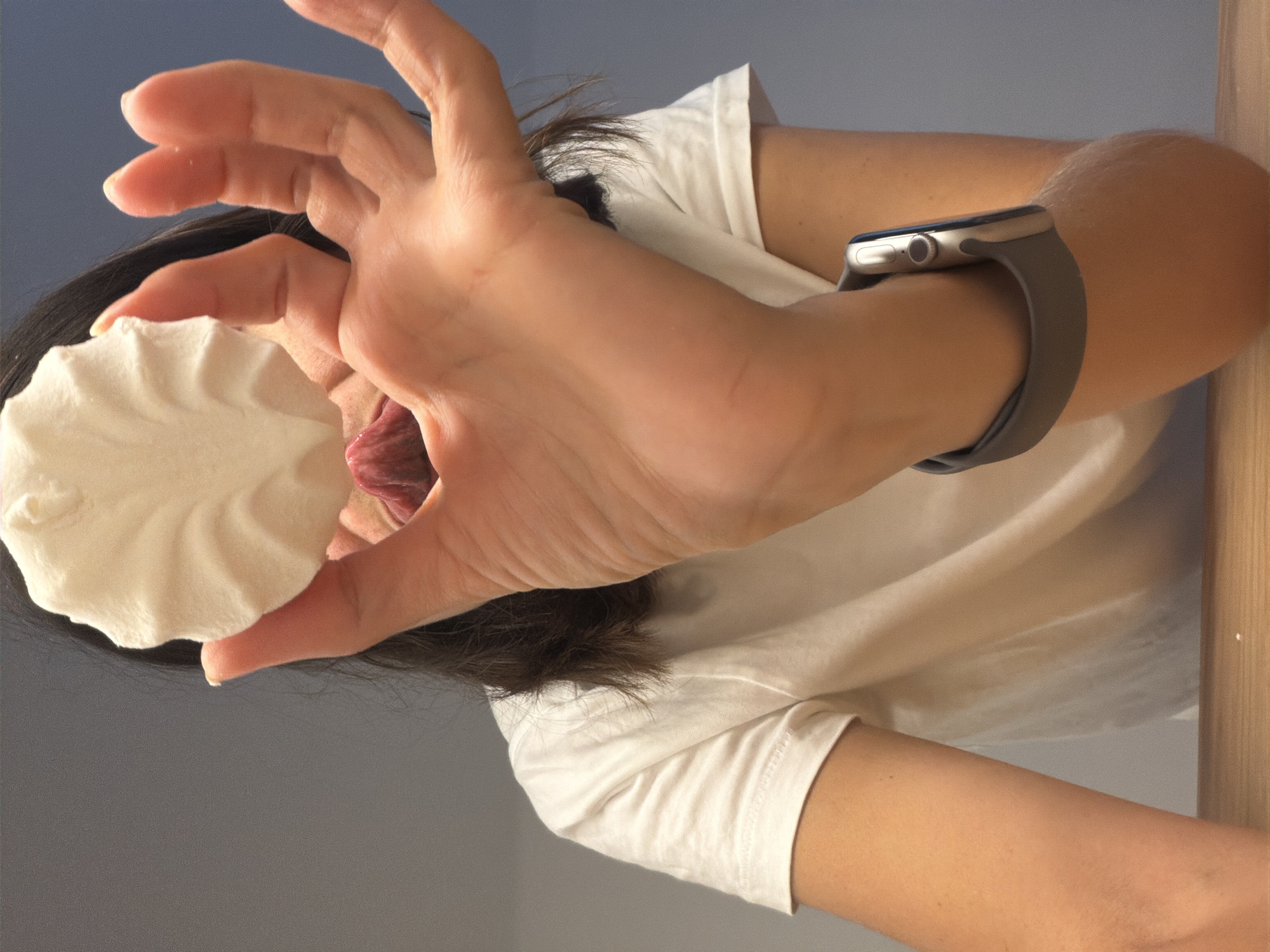 A woman is holding a white, round, meringue-like dessert in her left hand, close to her mouth, with her tongue extended as if to taste it. She is wearing a white t-shirt and an Apple watch with a gray band on her left wrist. The background consists of a gradient from gray to light blue. No specific location in Budva, Montenegro, is indicated.
