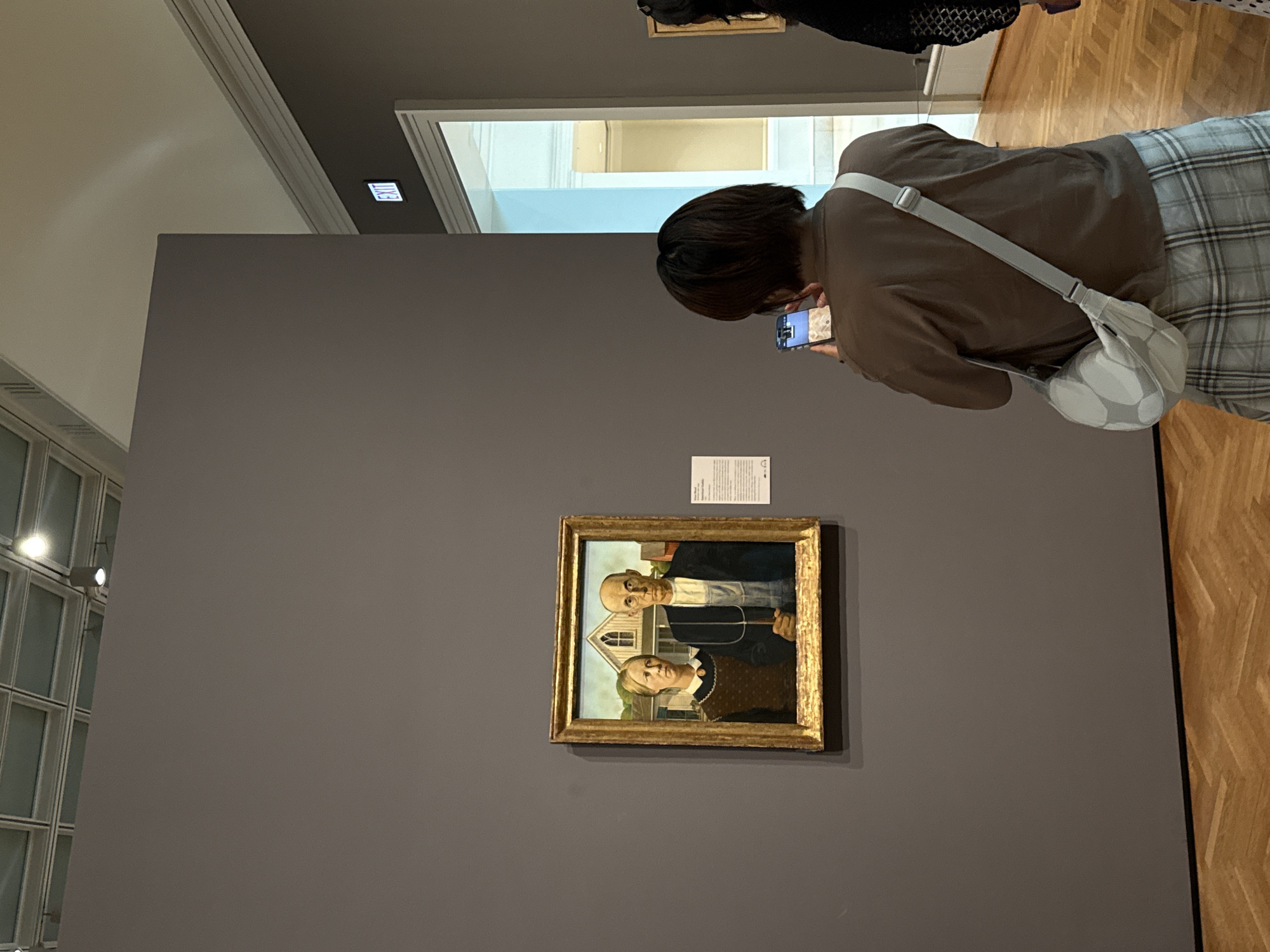 The image shows a section of a museum gallery. Prominently displayed on a large, dark gray wall is the iconic painting "American Gothic" by Grant Wood, housed in a gilded frame. To the right of the painting, a white placard with text is affixed to the wall. In the foreground and slightly to the right, a person with short, dark hair is seen from behind, wearing a brown long-sleeved shirt and a plaid skirt. This person is holding up a smartphone to capture a photograph of the painting. They are carrying a light-colored crossbody bag adorned with a pattern of light gray circles. The gallery floor is made of polished hardwood laid in a herringbone pattern. In the background, a bright, wide doorway with white trim is visible, leading to another section of the museum. An "EXIT" sign with illuminated green letters is positioned above the doorway. The ceiling features a grid of square panels with integrated lighting. The scene is characteristic of a museum exhibition space, likely in a major city like Chicago, known for its art institutions.