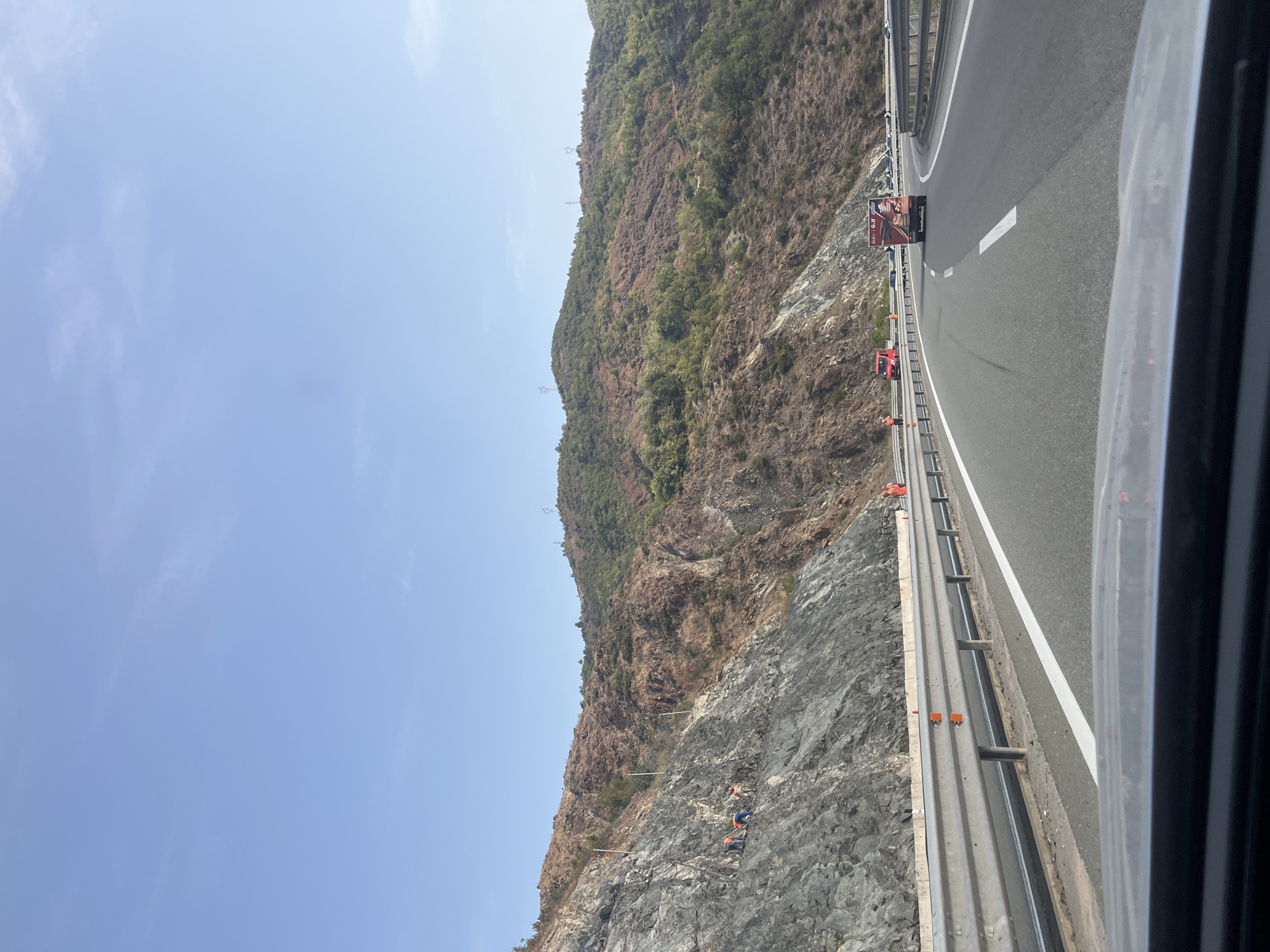 The image depicts a scenic highway scene in Rrëshen, Albania, with a mountainous backdrop. The highway is paved with asphalt and features clear lane markings. A metal guardrail runs along the side of the road, separating it from a steep, rocky embankment. Several individuals, dressed in orange safety vests, are visible working on the embankment, suggesting ongoing construction or maintenance.
The mountainous terrain is covered with sparse vegetation and features rocky outcrops, indicating a rugged environment. The sky above is clear and blue, suggesting fair weather. Various vehicles, including a red van and a truck with visible branding, are present on the highway, moving in the same direction as the viewer. The overall scene conveys a sense of activity and progress related to infrastructure development or repair in a natural, mountainous setting.