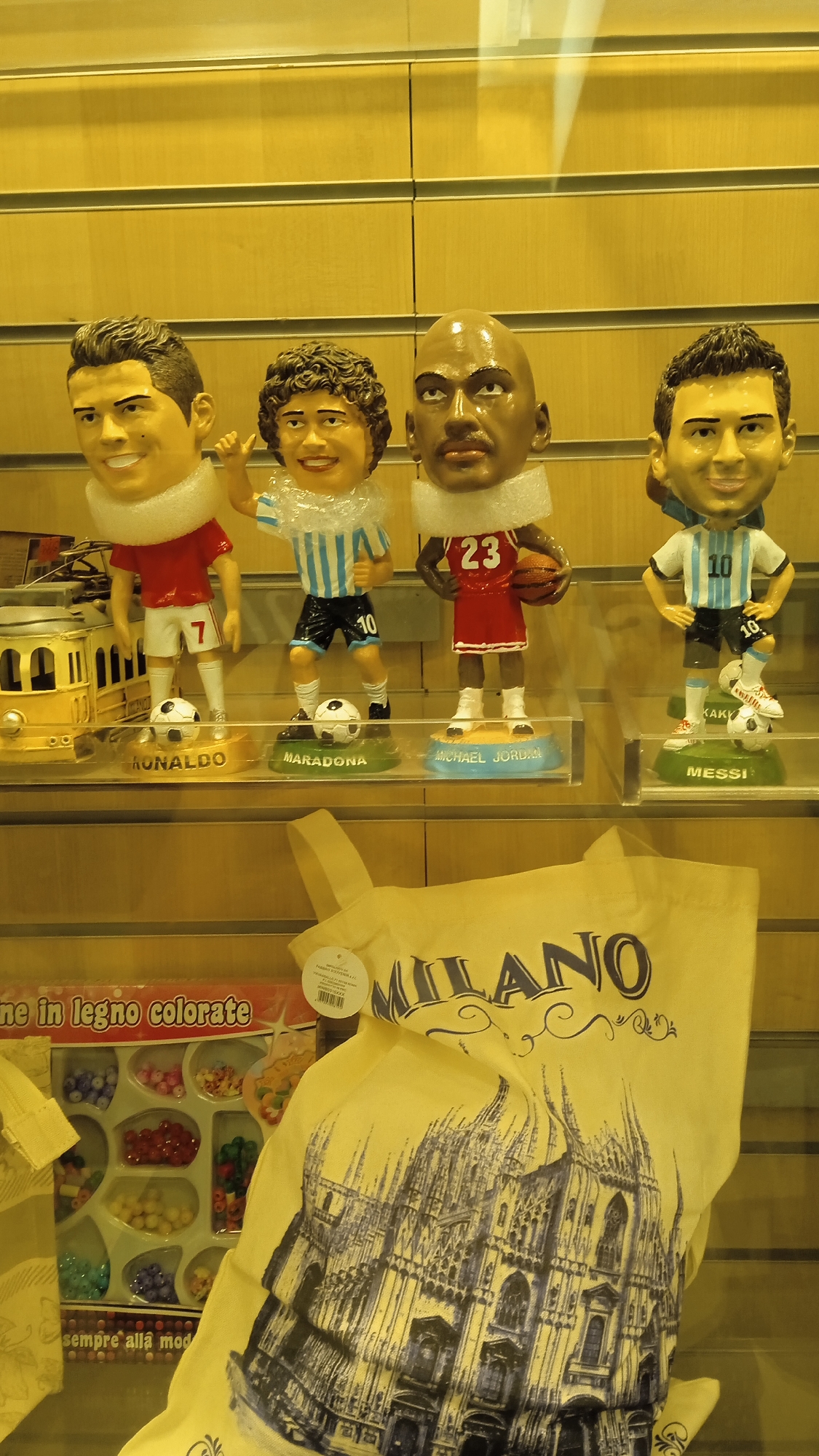 The image shows a display window with four bobblehead figures of famous athletes and a tote bag. From left to right, the figures are of Cristiano Ronaldo, Diego Maradona, Michael Jordan, and Lionel Messi. Each figure is standing on a base with their name written on it. Ronaldo is wearing a red soccer jersey with the number 7, Maradona is in a blue and white striped soccer jersey with the number 10, Jordan is in a red basketball jersey with the number 23 and holding a basketball, and Messi is in a blue and white striped soccer jersey with the number 10.
To the left of the figures, there is a toy yellow tram. Below the figures, there is a box of colorful beads with the text "in legno colorate" and "sempre alla moda" which translates to "wooden colored" and "always in fashion".
In the foreground, a yellow tote bag is draped over the display shelf. The bag has the word "MILANO" printed on it in large, decorative letters, and a black and white sketch of the Milan Cathedral (Duomo di Milano). The background consists of horizontally slatted wooden panels. The overall impression is that of a retail display, likely in Milan, Italy, selling souvenirs related to sports and the city.
