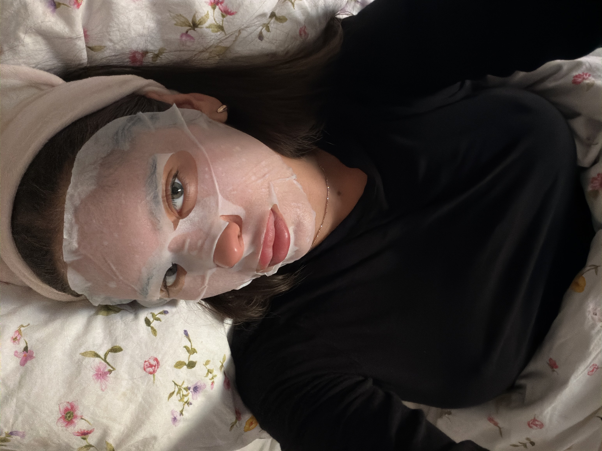 A woman is lying down on a bed with floral patterned bedding. She is wearing a facial mask and a light pink headband. She has dark hair and is wearing a long-sleeved black shirt. A gold necklace is visible around her neck. The photo appears to be taken from an overhead perspective. The city of Krasnoyarsk, Russia is not discernable from this image.