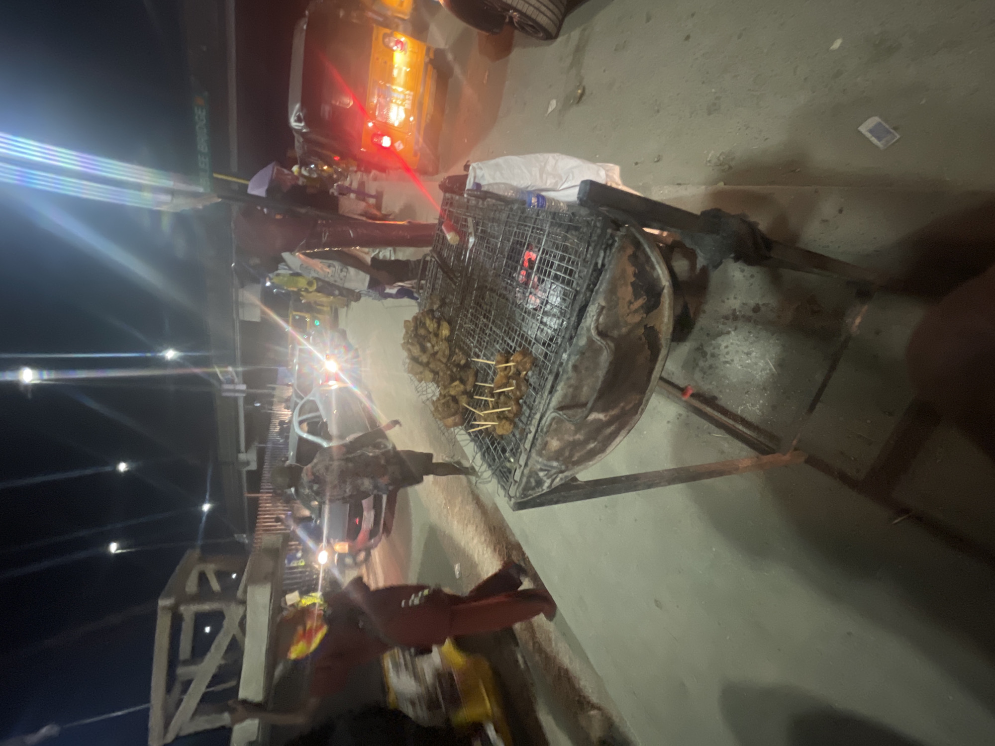 The image shows a street food scene at night, likely in Lagos, Nigeria, based on the "3rd Mainland Bridge" sign visible in the background. A metal grill laden with skewers of meat (possibly suya or grilled beef) is positioned on a metal stand. The grill is blackened from use and contains visible embers. The surrounding environment includes a concrete road surface, street lights, and passing vehicles, including a yellow "keke" or tricycle taxi. Several people are visible around the grill, seemingly engaging in the preparation or sale of the street food. The overall lighting is dim, with artificial light sources creating a somewhat hazy ambiance.
