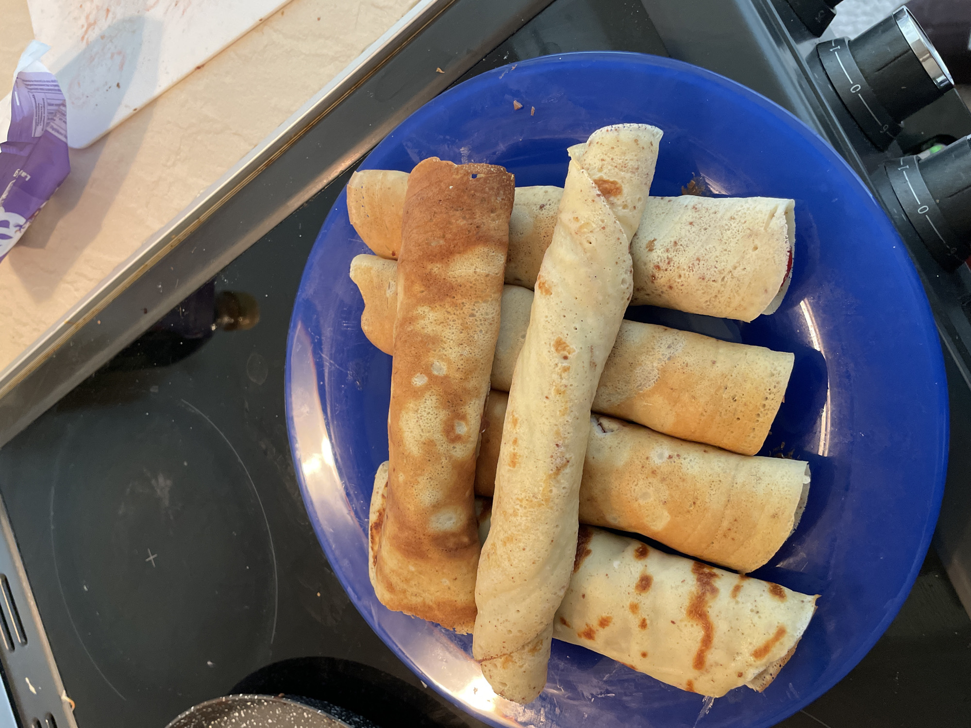 A bright blue plate filled with rolled pancakes rests on a black stovetop. The pancakes vary in color from golden brown to lighter beige with speckles, suggesting different cooking times or ingredients. The plate is full, with the pancakes stacked on top of each other. To the left of the plate, a portion of a countertop is visible, along with a wrapper, likely from a food product. To the right, the stovetop extends, showing the controls for the burners. The setting is indoors, likely a kitchen. It is impossible to determine if the location is in Zlatograd, Bulgaria, or any specific city based on the provided image.