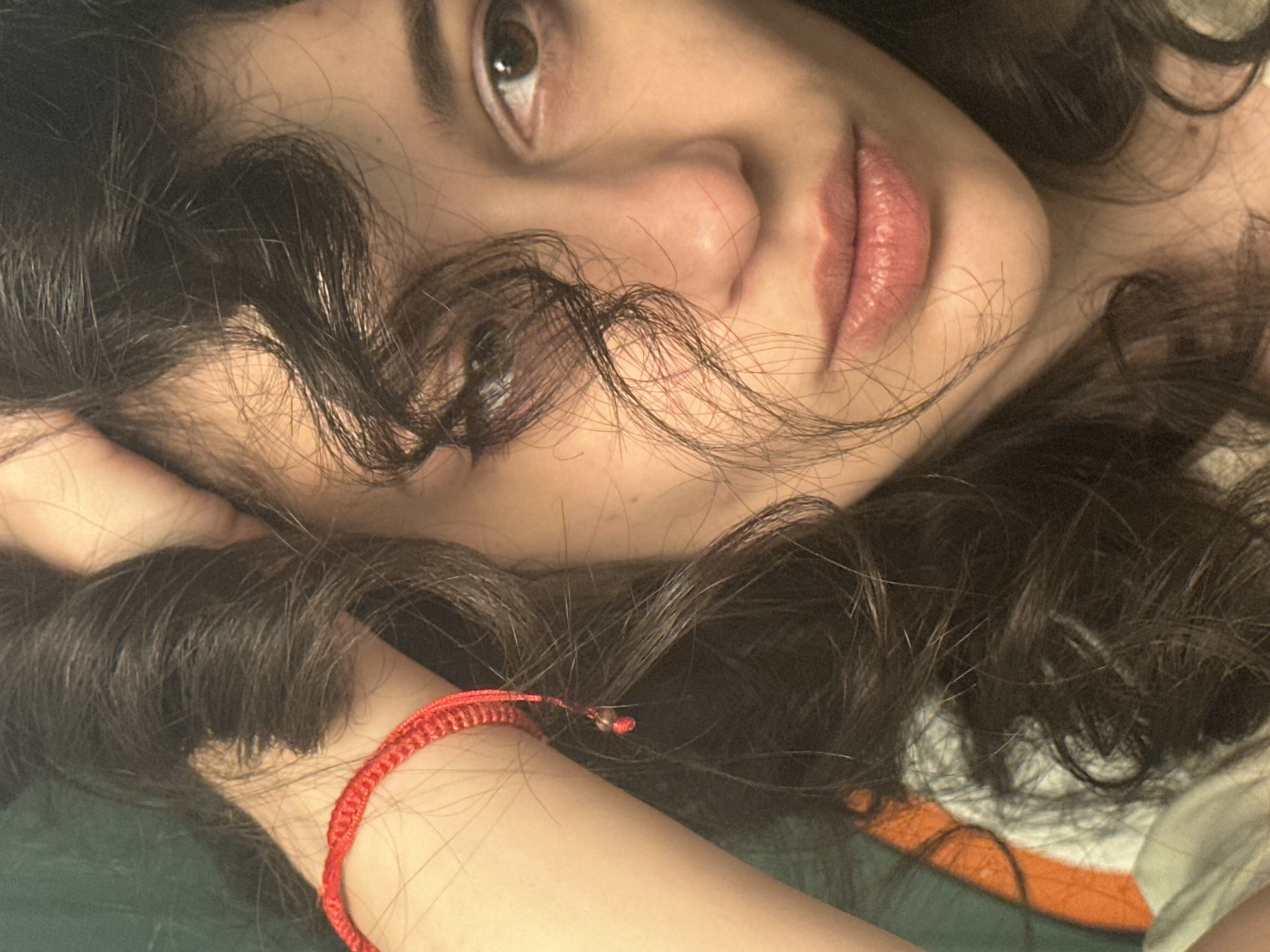 The image is a close-up, portrait-style photograph of a young woman's face and upper body. She has dark, curly hair that is partially obscuring her face. Her left hand is raised and touching her hair. She is wearing a red braided bracelet on her left wrist. Her clothing is partially visible and appears to be a light-colored t-shirt with orange and white stripes. The background is a dark green wall. The lighting is soft and natural, suggesting an indoor setting. There is no information in the image to indicate the location within the city of Sofia, Bulgaria.