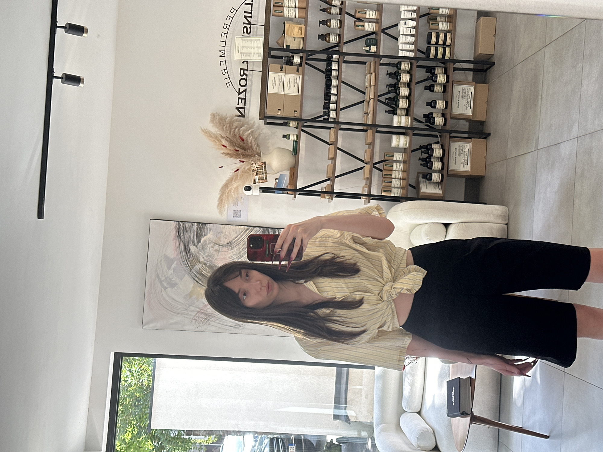 The image shows a young woman taking a selfie in what appears to be a retail setting. She is holding a red cell phone. She is wearing a yellow striped shirt tied at the waist and black shorts. She has long dark hair and fair skin.
Behind her is a shelf stocked with products in packaging that is suggestive of cosmetics or personal care items. There is an abstract painting hanging on the wall. There are white sofas and a small side table. There's a sign that reads "Bebe Boutique" backwards, suggesting this is a mirrored image. Natural light comes through a window.
The location is possibly in the Vracar Urban Municipality, Serbia, based on the assumption that the retail establishment is in that area. However, this location is solely based on the prompt parameters.