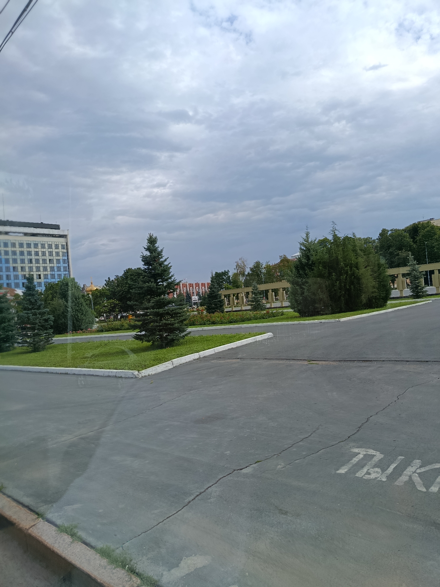 A side view, captured from inside a vehicle, shows a paved area with a white-lined border and a curb. The pavement is cracked in places and has some water stains. In the background, a meticulously kept lawn features several evergreen trees, including a prominent fir tree. Beyond the lawn, a building with a golden dome is visible, along with a row of buildings with arched facades. The sky is overcast with clouds. The image appears to be taken in Saratov, Russia, specifically near the Saratov State University building, indicated by the architectural style and landscaping reminiscent of the university grounds. A partial inscription in Russian, "ТЫК," is visible on the pavement in white paint.