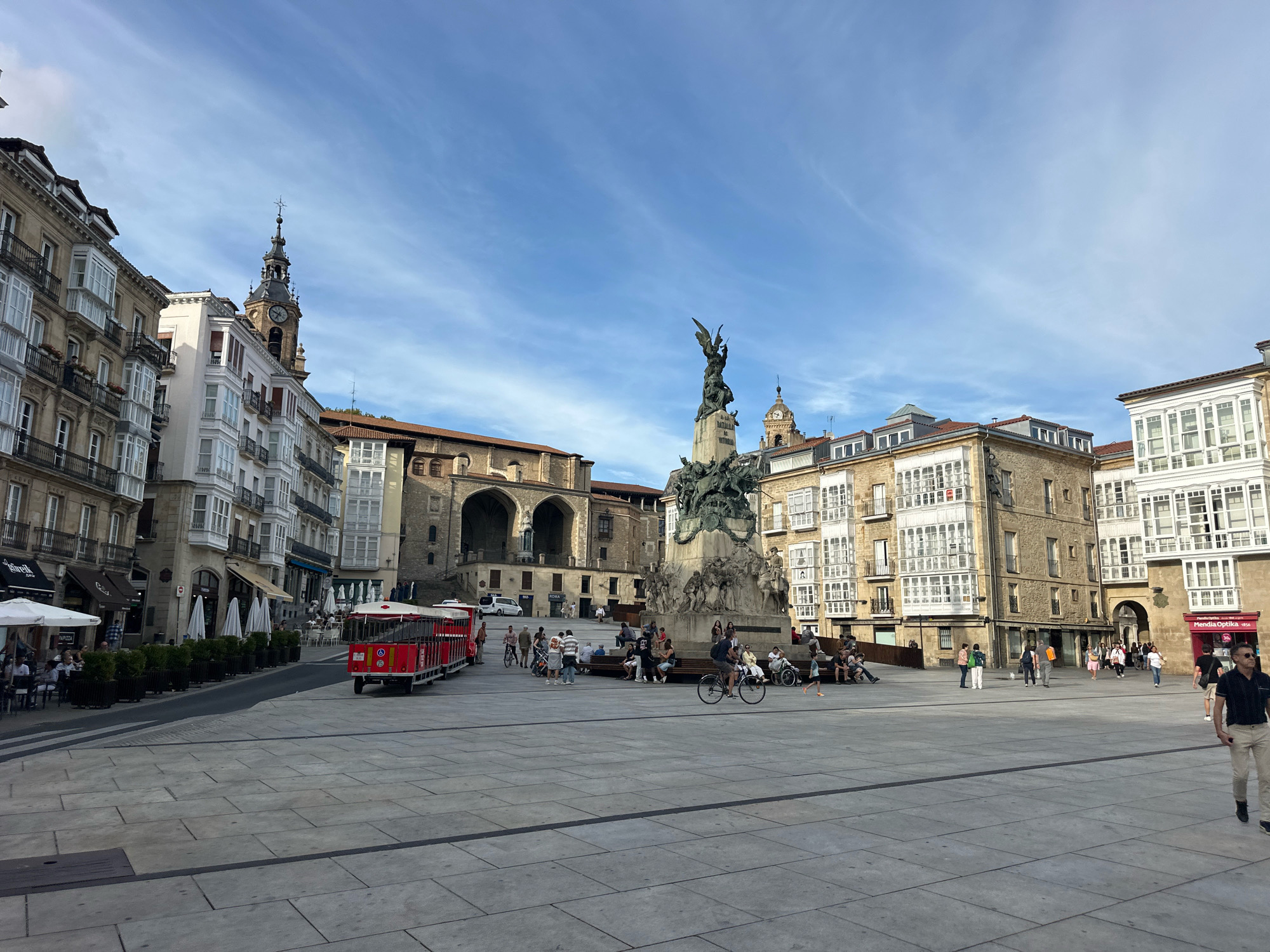 The image depicts a wide, paved public square in Vitoria-Gasteiz, Spain, likely Plaza de la Virgen Blanca. The square is lined with multi-story buildings featuring traditional European architecture, characterized by balconies with white-framed windows. In the background, a tall clock tower with a spire rises above the buildings. Dominating the center of the square is a large, elaborate monument featuring a sculpture of winged figures and equestrian statues.
Several people are present in the square, engaging in various activities. Some are walking, others are sitting on benches around the monument or at outdoor cafe tables. A red tourist train with open carriages is visible on the left side of the square, appearing to be stopped or moving slowly. A man on a bicycle is seen in the mid-ground, and a child is running in the right mid-ground. The sky above is blue with scattered clouds. The overall scene conveys a lively, urban atmosphere with elements of historical architecture and public recreation.