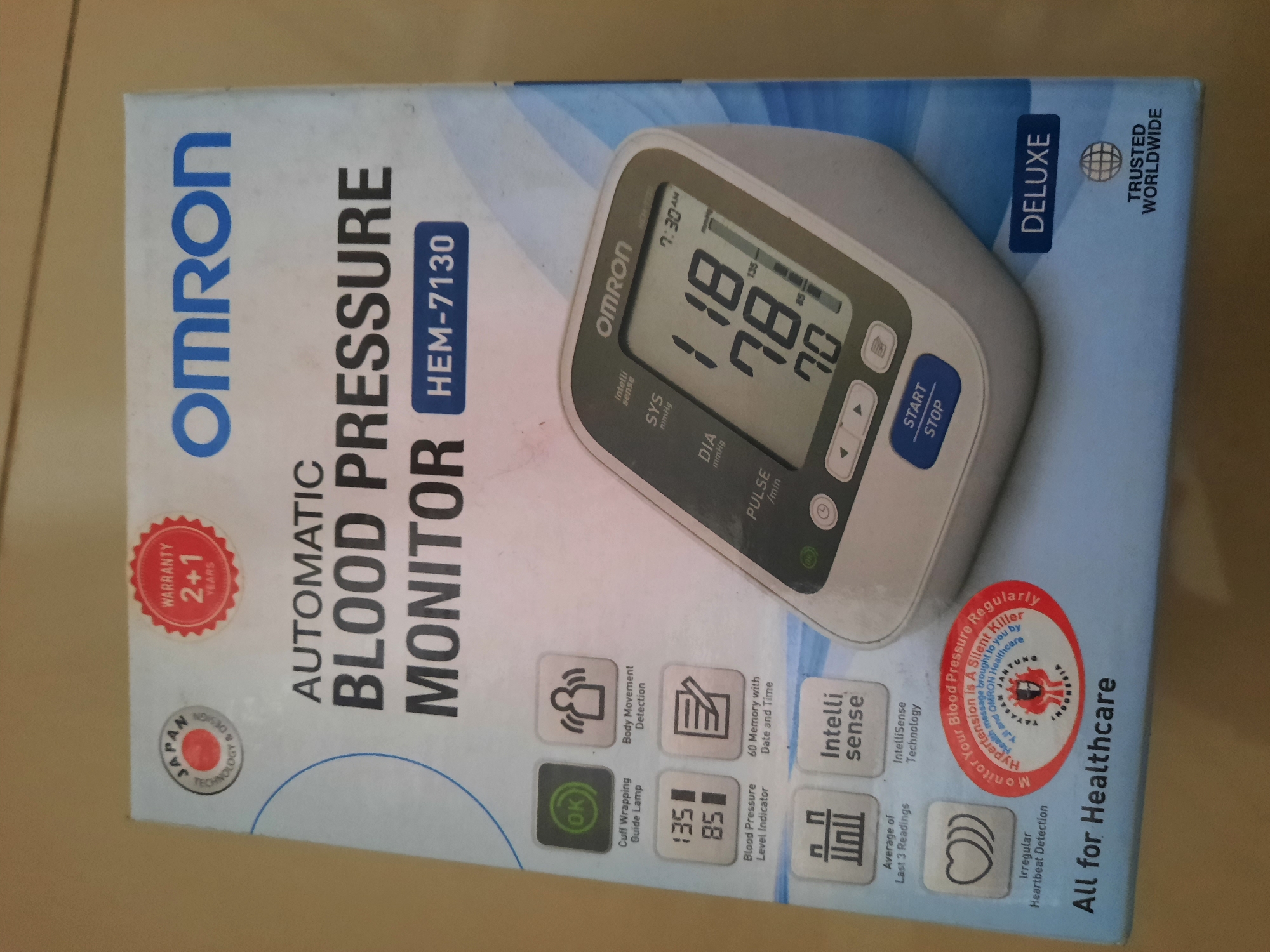 The image shows the front of a box for an OMRON Automatic Blood Pressure Monitor, model HEM-7130. The box is primarily white with blue and red accents. Prominently displayed are the OMRON logo, "AUTOMATIC BLOOD PRESSURE MONITOR", and "HEM-7130". Various icons indicate features such as "Cuff Wrapping Guide Lamp", "Body Movement Detection", "Blood Pressure Level Indicator", "60 Memory with Date and Time", "Average of Last 3 Readings", and "Irregular Heartbeat Detection". A digital display of a blood pressure monitor is visible, showing readings of 118/78 with a pulse of 70. A circular red sticker reads "Monitor Your Blood Pressure Regularly", with text about hypertension being a silent killer. Below the main product details, it says "All for Healthcare" and "DELUXE TRUSTED WORLDWIDE". The box appears to be photographed in a dimly lit indoor environment with a plain, textured background, possibly a wall or table. There are no people or discernible geographical markers within the image that would indicate a specific location in Lhokseumawe, Indonesia.