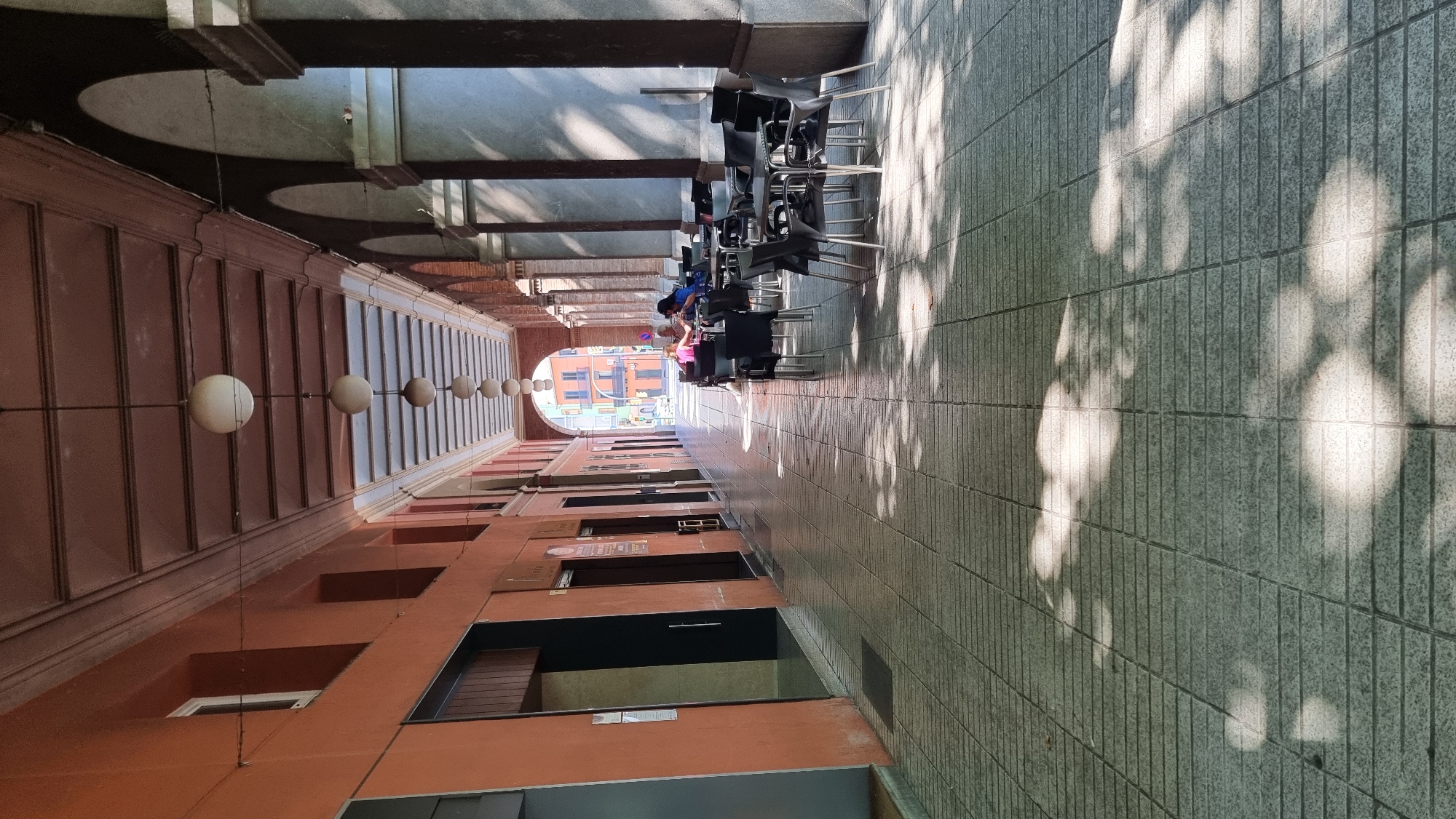 The image shows a long, covered walkway with arches on the right side, supported by columns. The walkway is paved with gray tiles and has a tiled ceiling. Several spherical lights hang from the ceiling, spaced evenly along the walkway. On the left, the wall is painted orange, with several dark doorways. On the right, tables and chairs are set up, suggesting an outdoor cafe or restaurant area. Two people are seated at one of the tables, with one person looking towards the other. Sunlight filters through the arches, casting shadows on the floor. In the distance, buildings are visible at the end of the walkway, including a colorful orange and yellow building. The scene appears to be in a public area of a Spanish city, possibly Olot, given the architectural style.